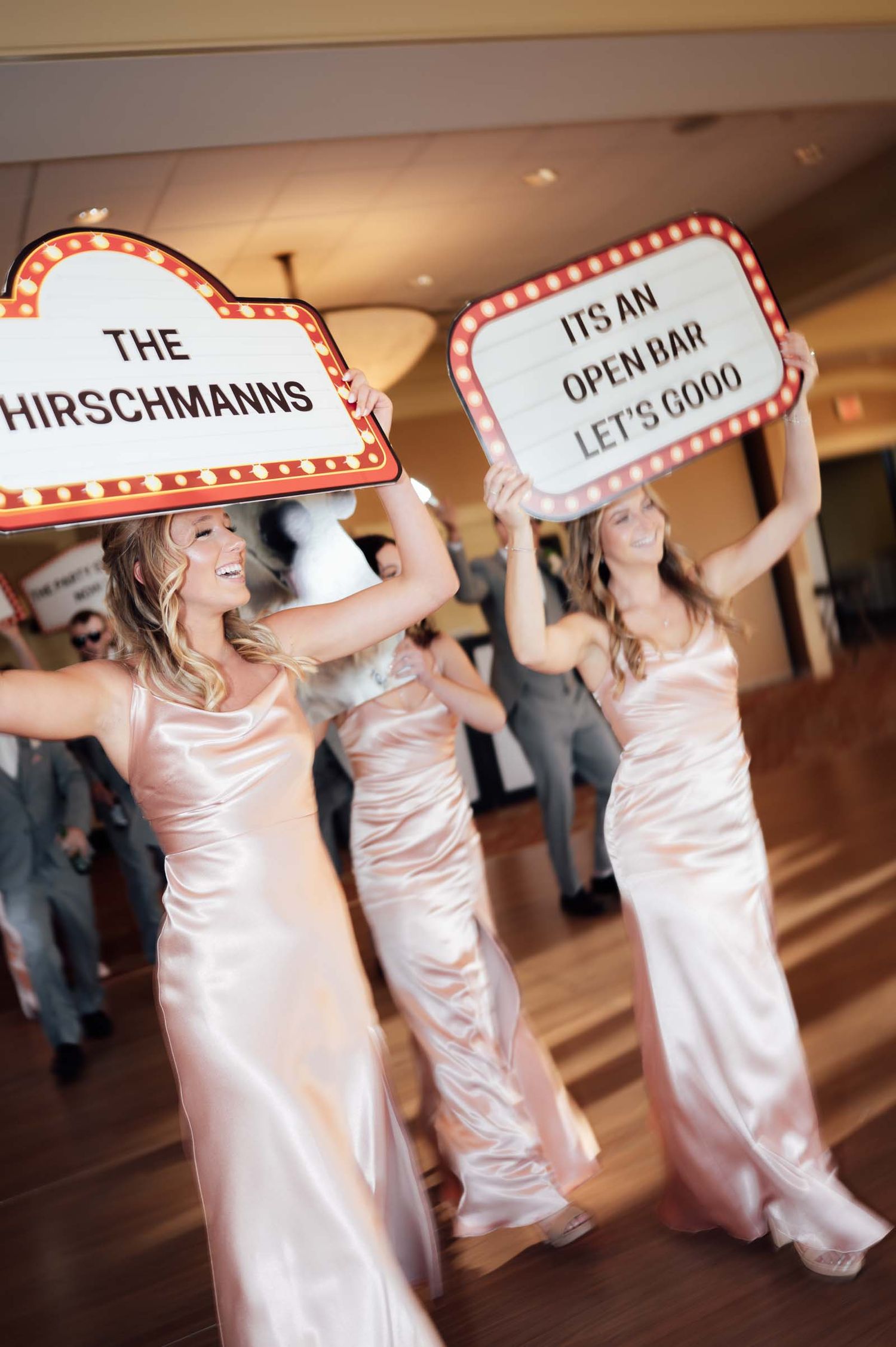 Bridesmaids in pink satin gowns carrying marquee signs announcing the Hirschmanns and open bar.