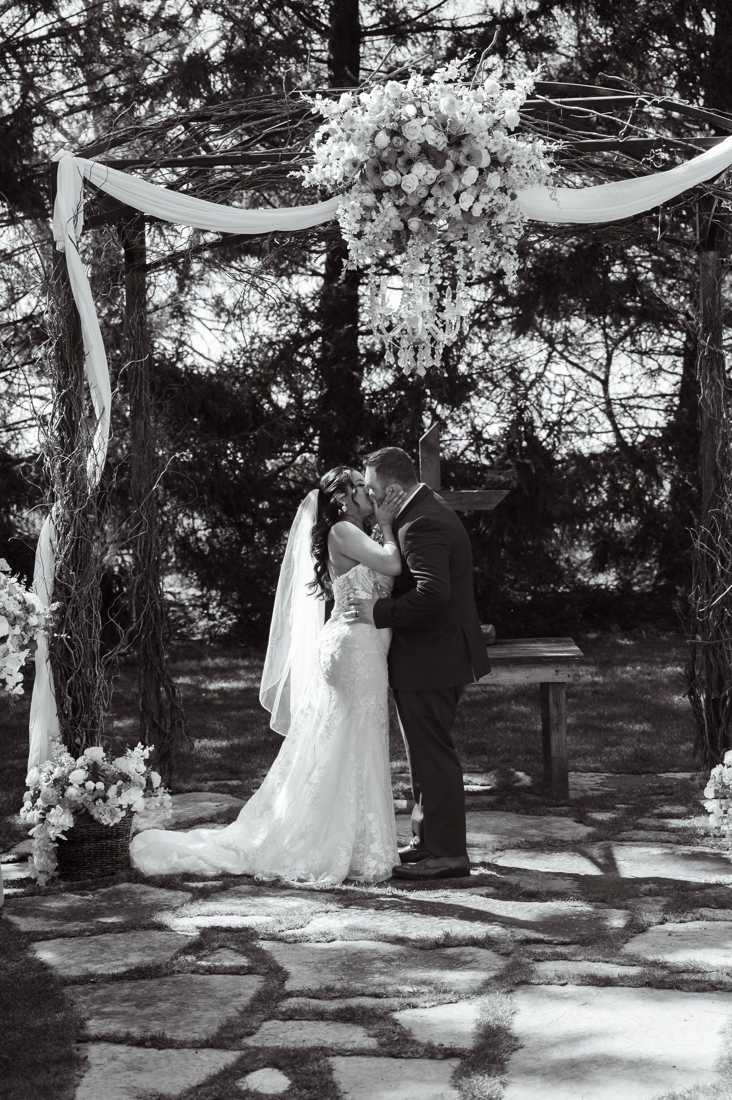 Black and white wedding photo of couple sharing a kiss under a floral arch decorated with white fabric in a wooded setting.