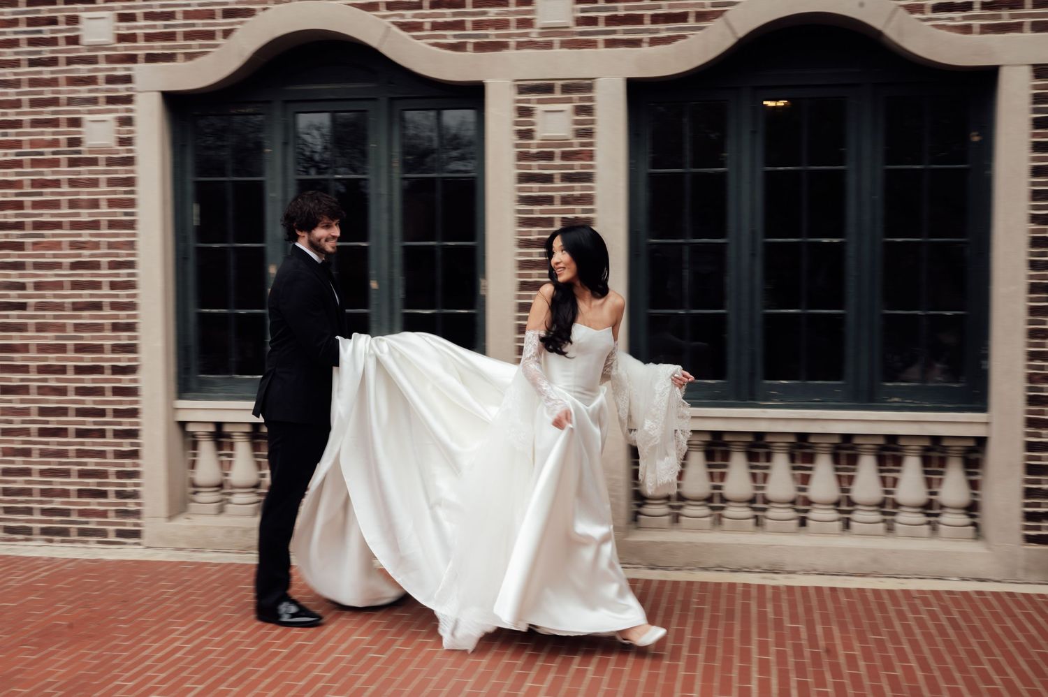 Bride in elegant white gown twirling with groom in black suit outside brick building with arched windows.