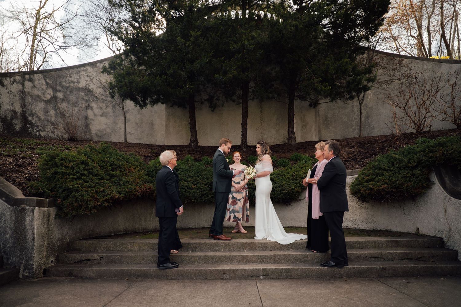 Small outdoor wedding ceremony on stone steps with bride, groom, officiant, and two guests surrounded by greenery.