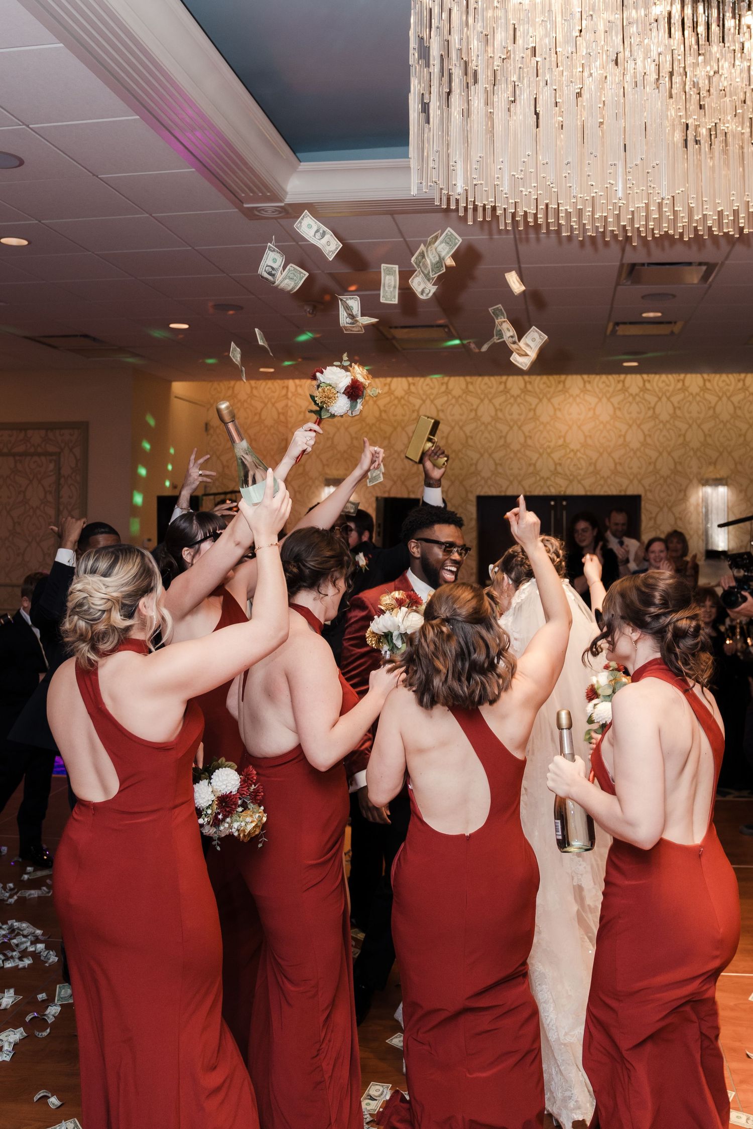 Bridesmaids in red dresses dancing and celebrating together under a crystal chandelier at a wedding reception.