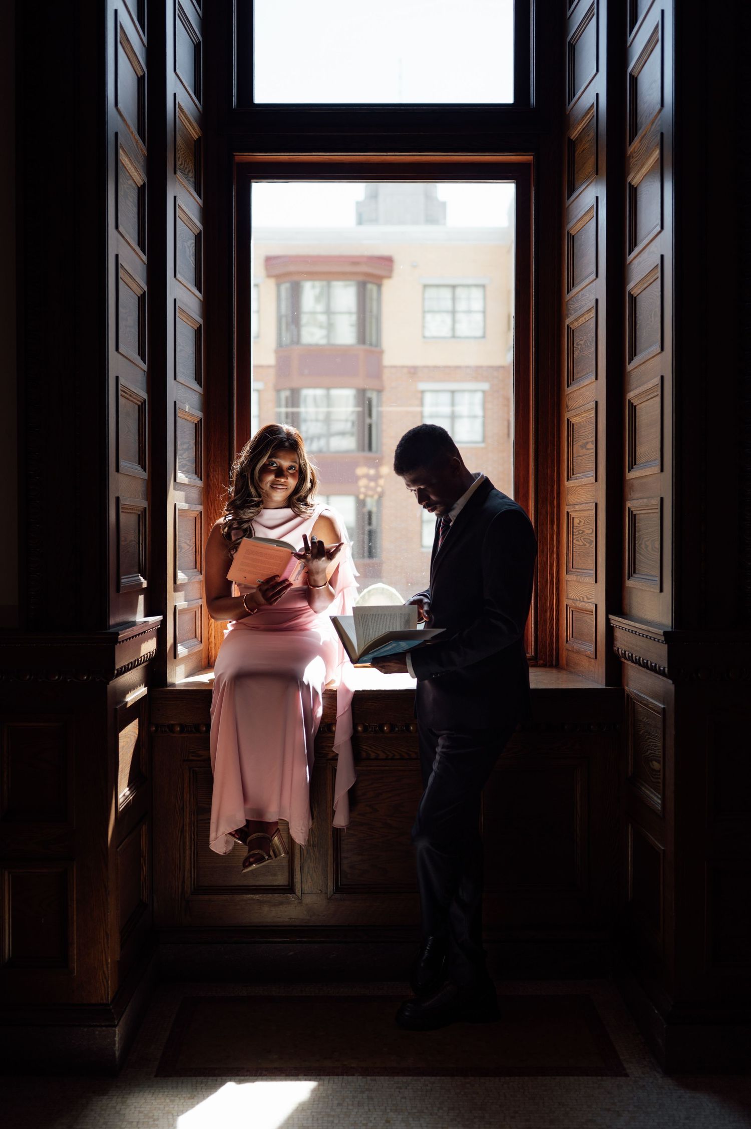 A couple reads books together by a sunlit window in an elegant dark wood hallway, woman in pink dress, man in suit.