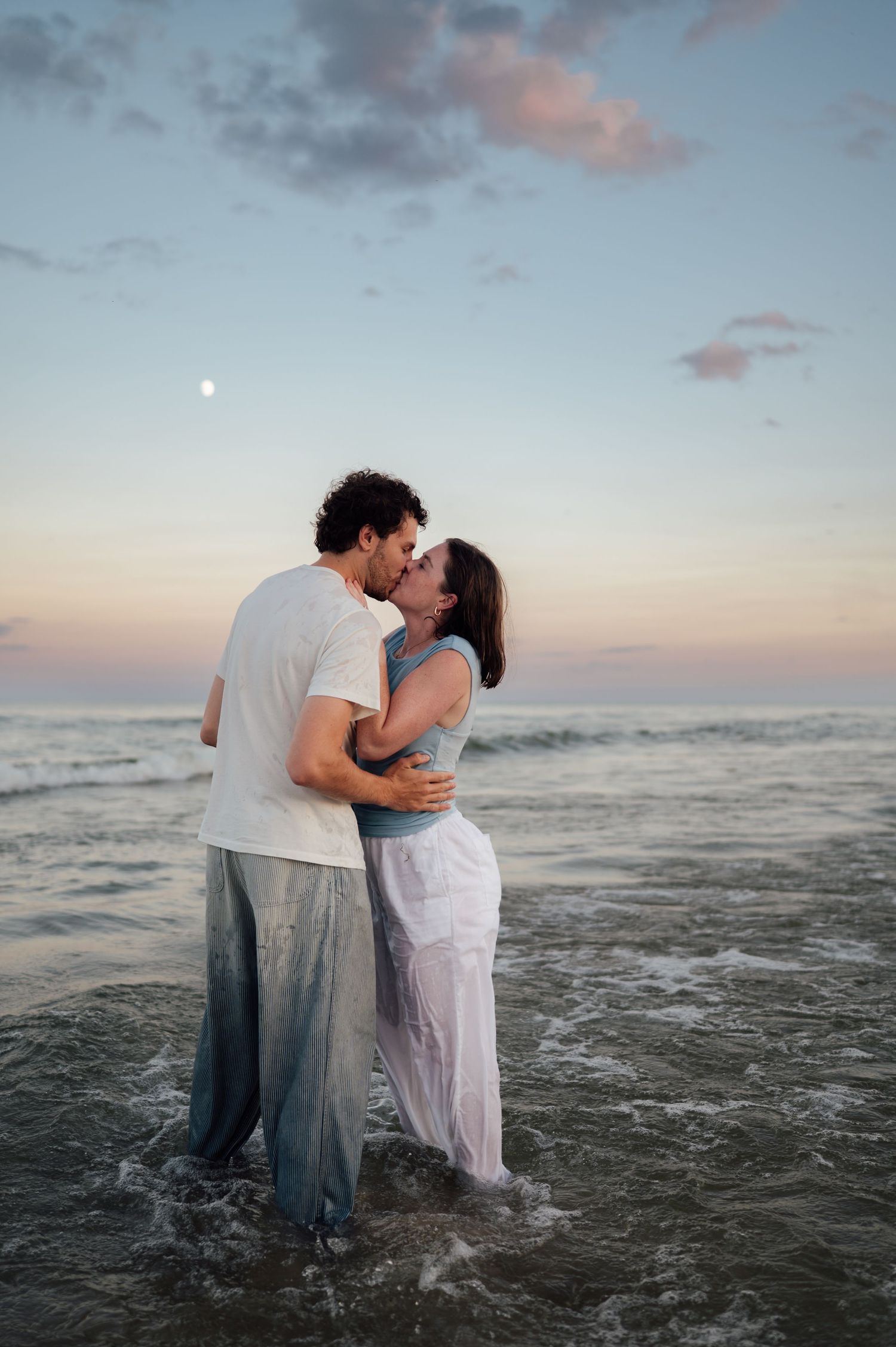 Romantic couple kissing at sunset on the beach, standing in ocean waves with moon visible in pastel sky.