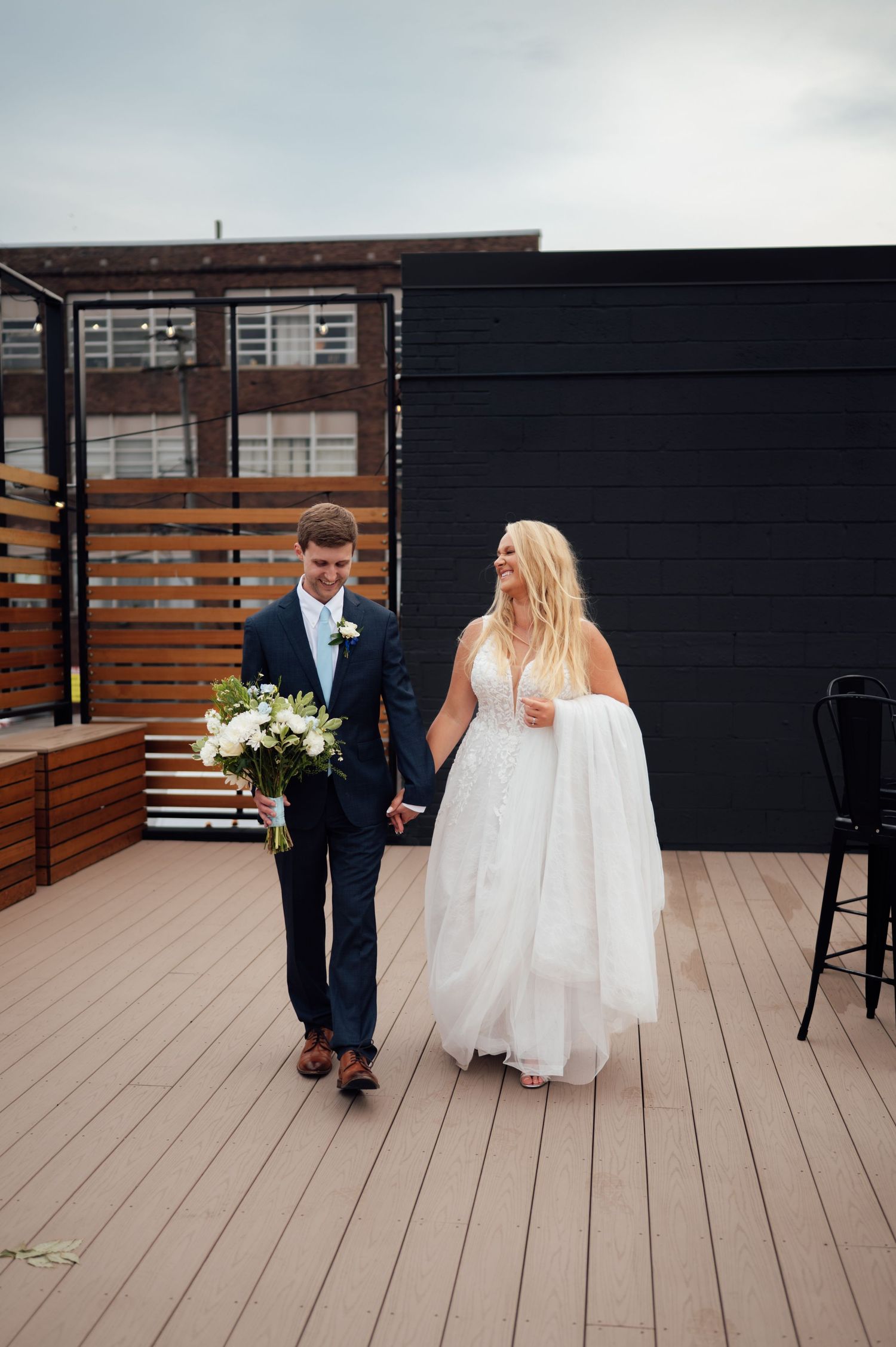 Bride and groom walk hand-in-hand on a rooftop deck, bride in white gown holding dress train, groom in navy suit with bouquet.