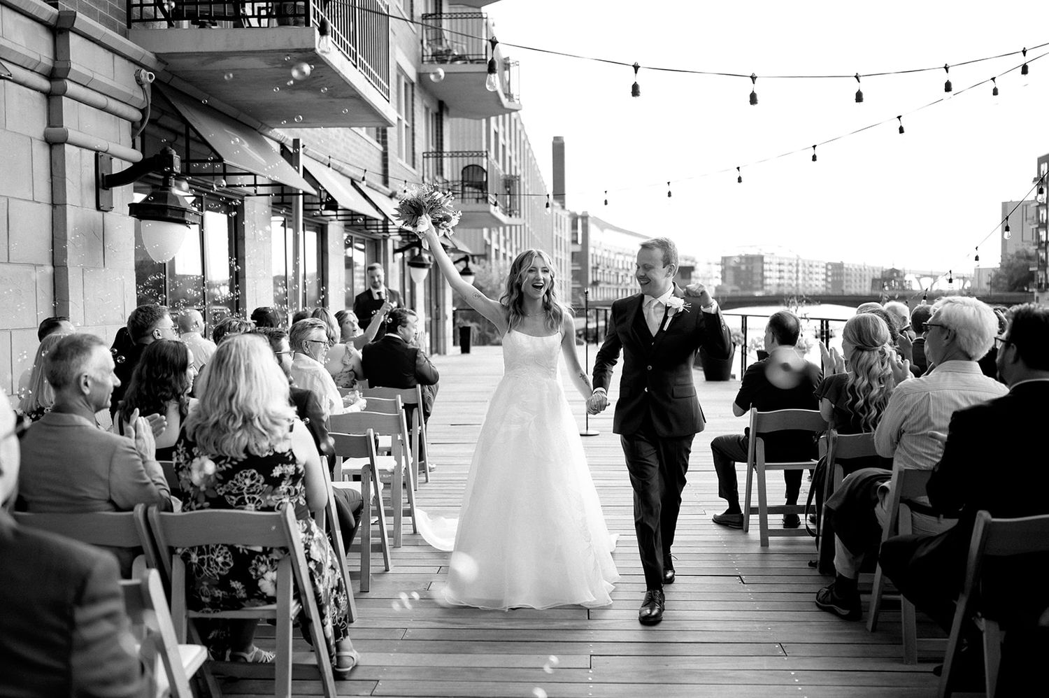 Black and white photo of bride and groom walking down outdoor waterfront wedding ceremony aisle.