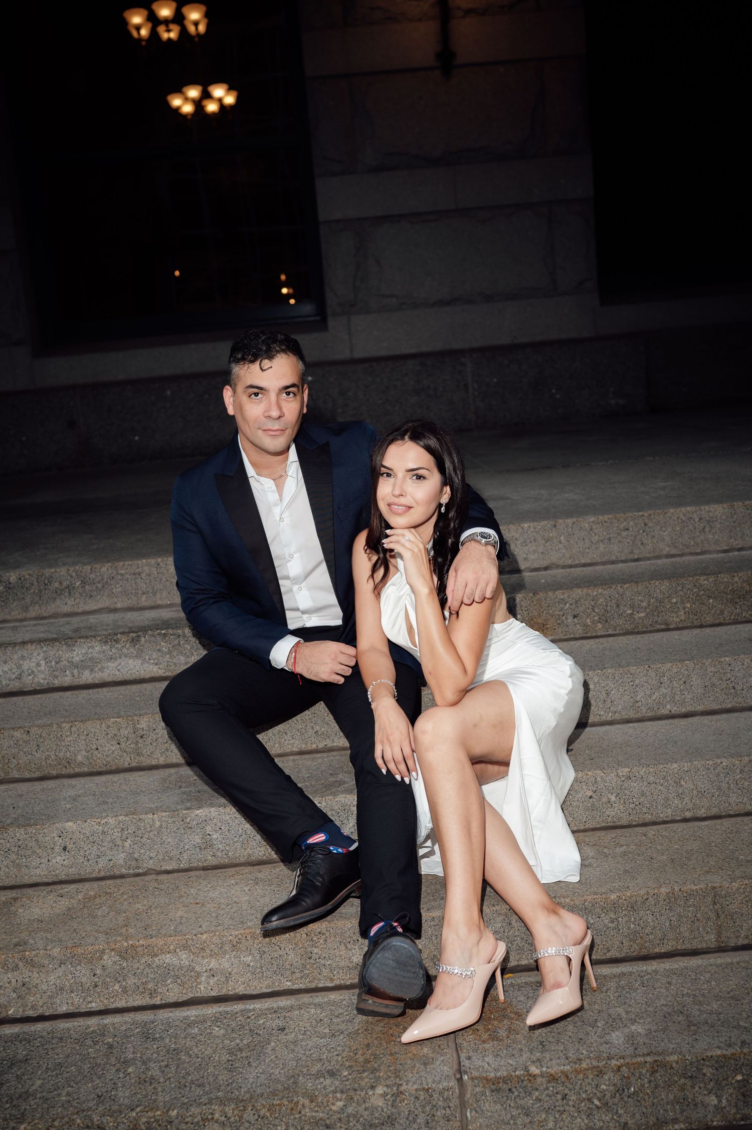 Elegant couple sitting on stone steps at night, woman in white dress and man in navy suit posing together.
