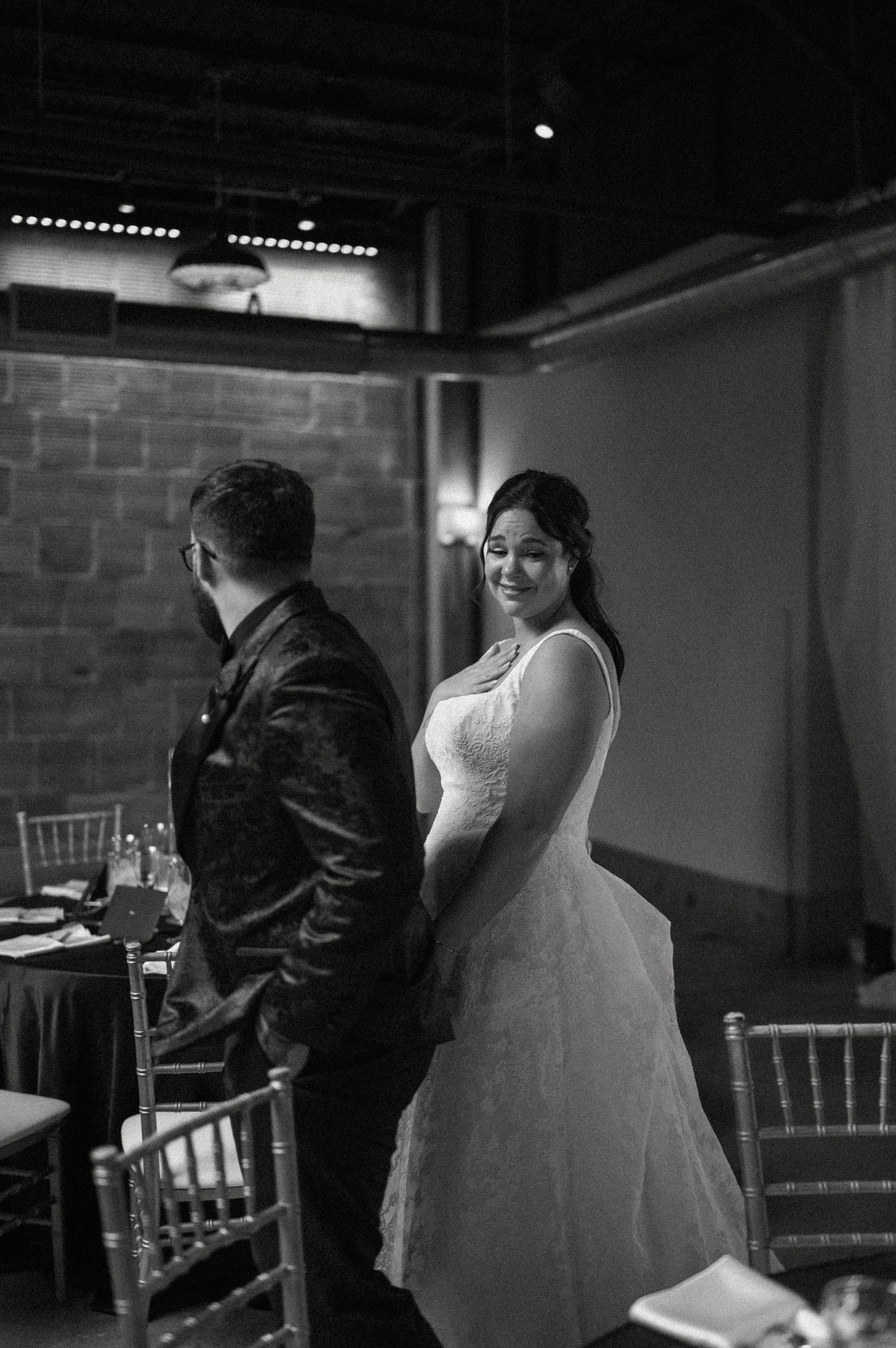 Black and white wedding photo of smiling bride in lace gown glancing back at groom in elegant reception venue.
