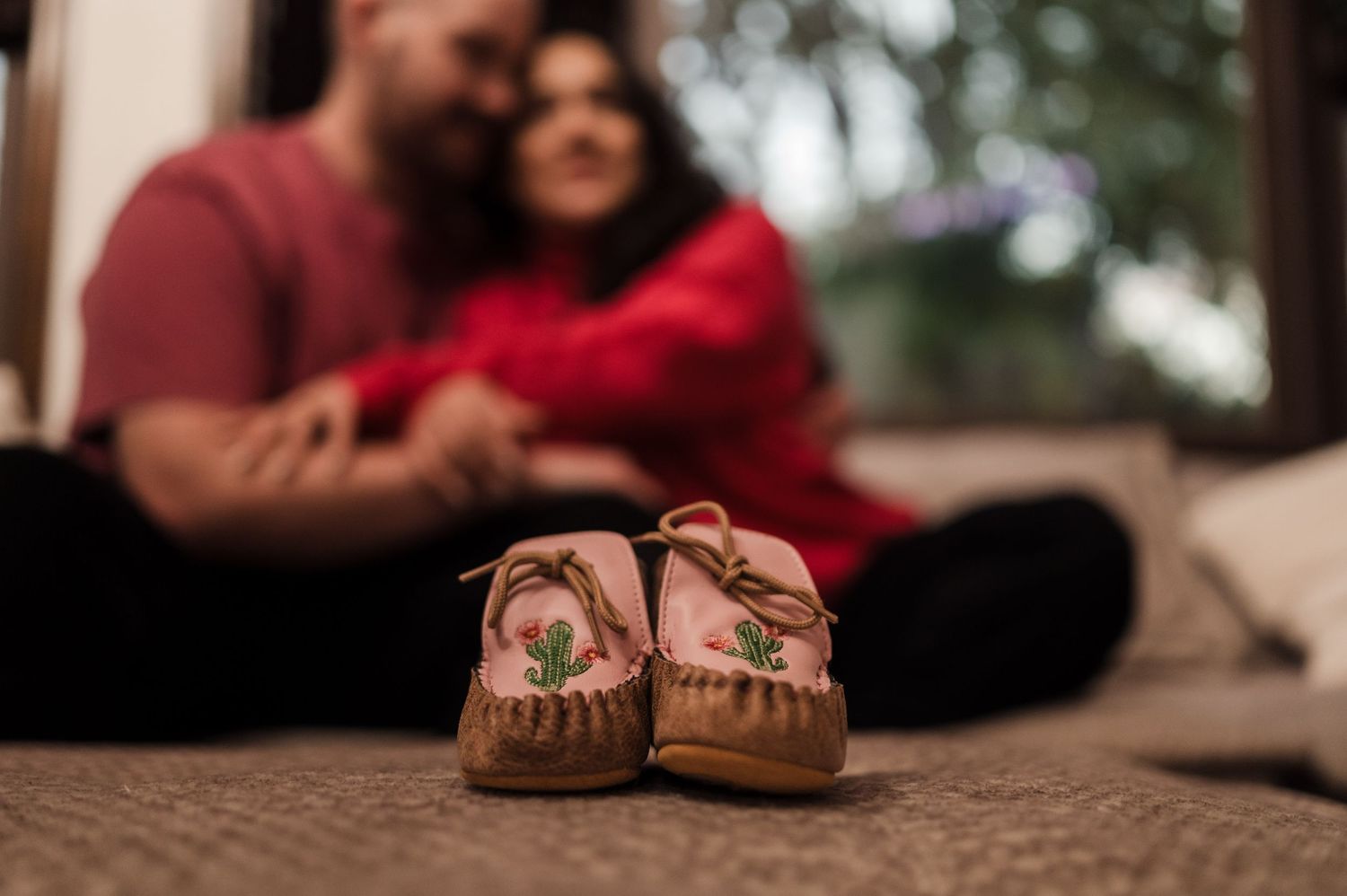 Tiny cactus baby moccasins in foreground with expecting couple sitting together in soft background.