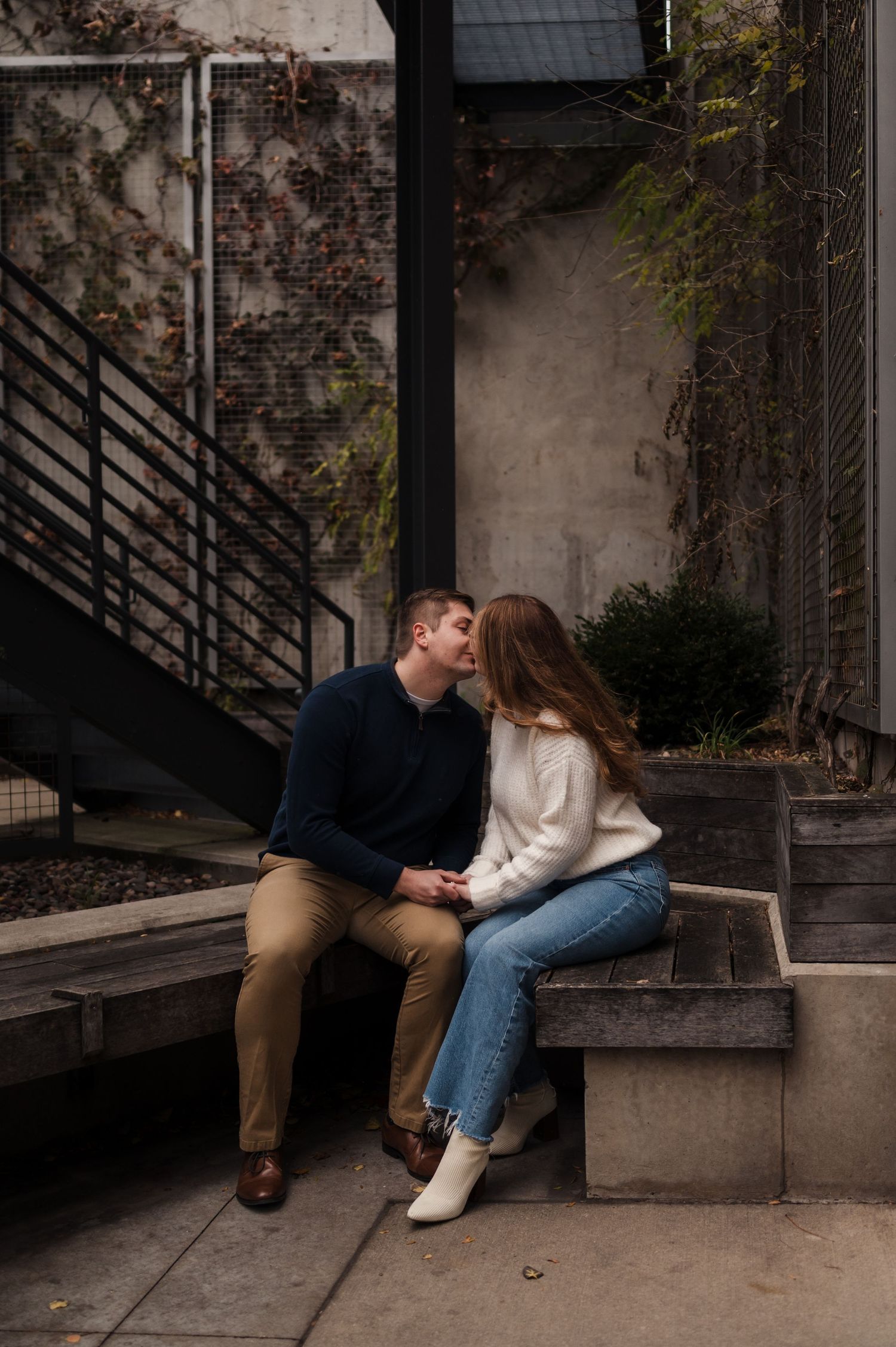 Couple sharing a kiss while seated on outdoor concrete steps near an urban staircase.