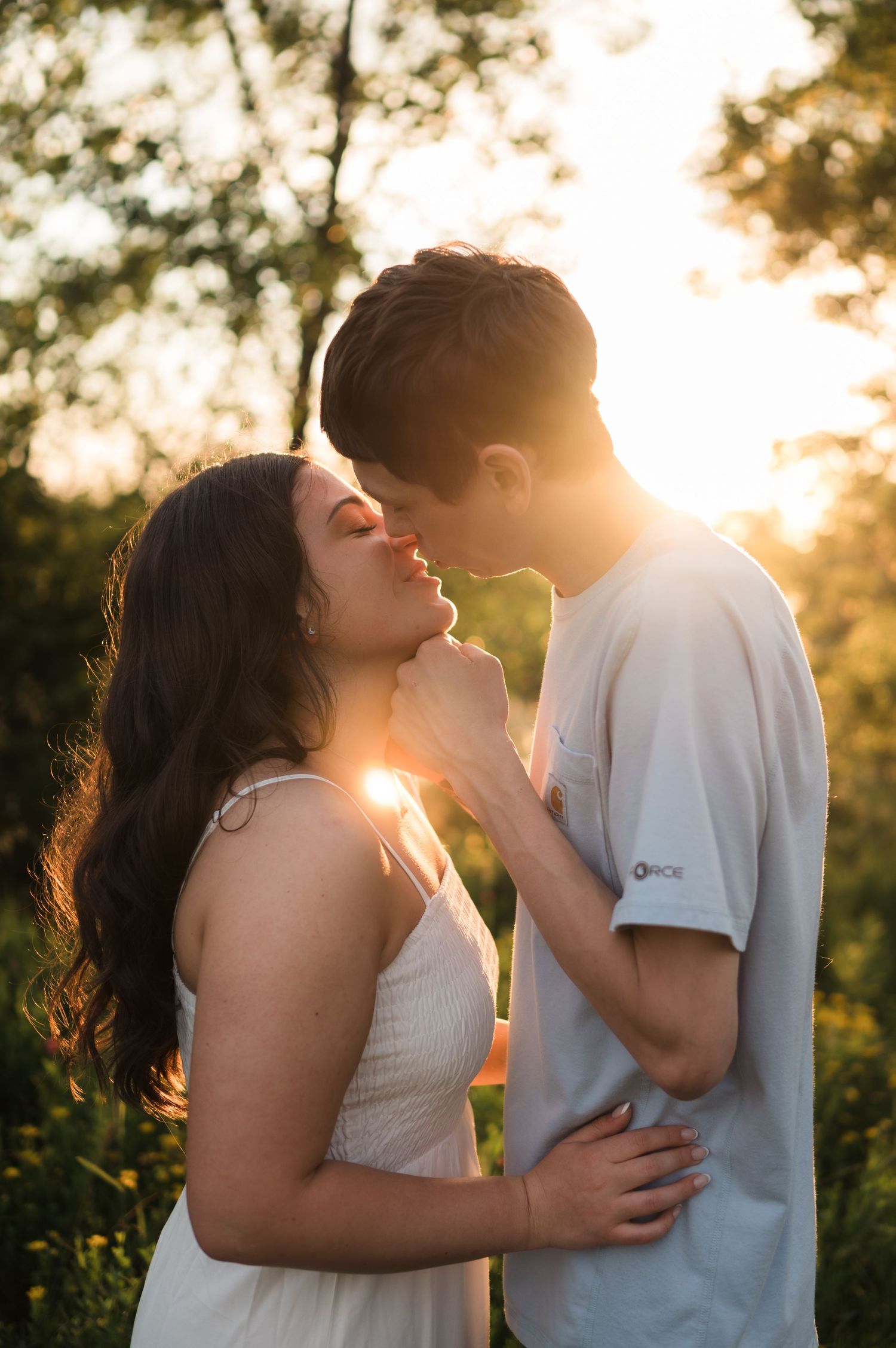 Couple about to kiss in a sun-drenched field with warm golden backlight filtering through surrounding trees.