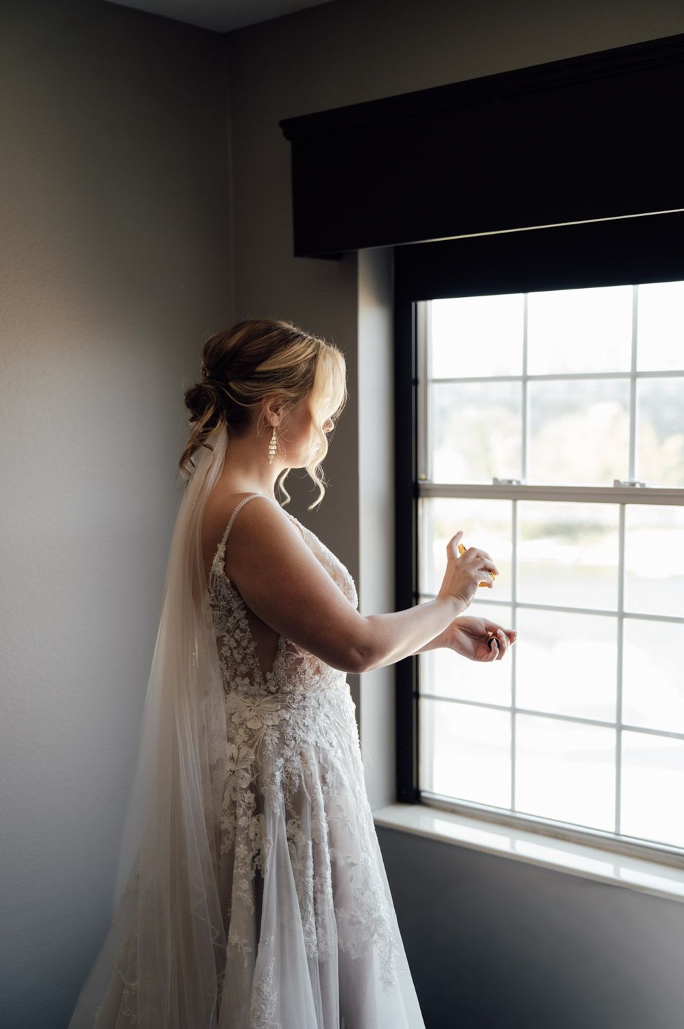 Elegant bride in lace wedding gown and veil stands by window applying perfume before ceremony.
