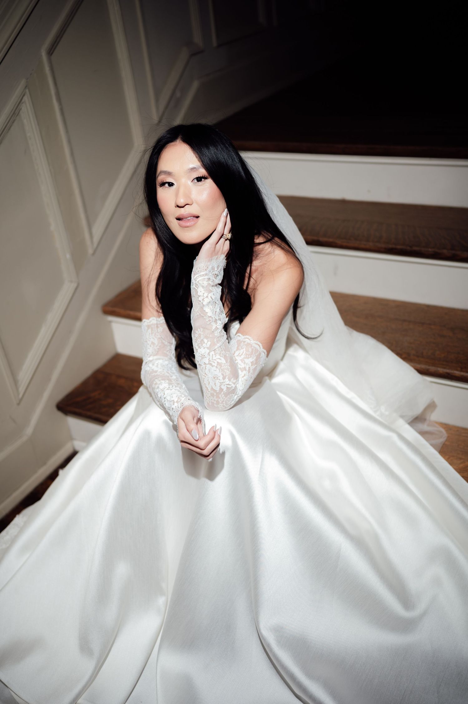 Elegant bride in white satin ballgown and lace gloves poses gracefully on wooden staircase.
