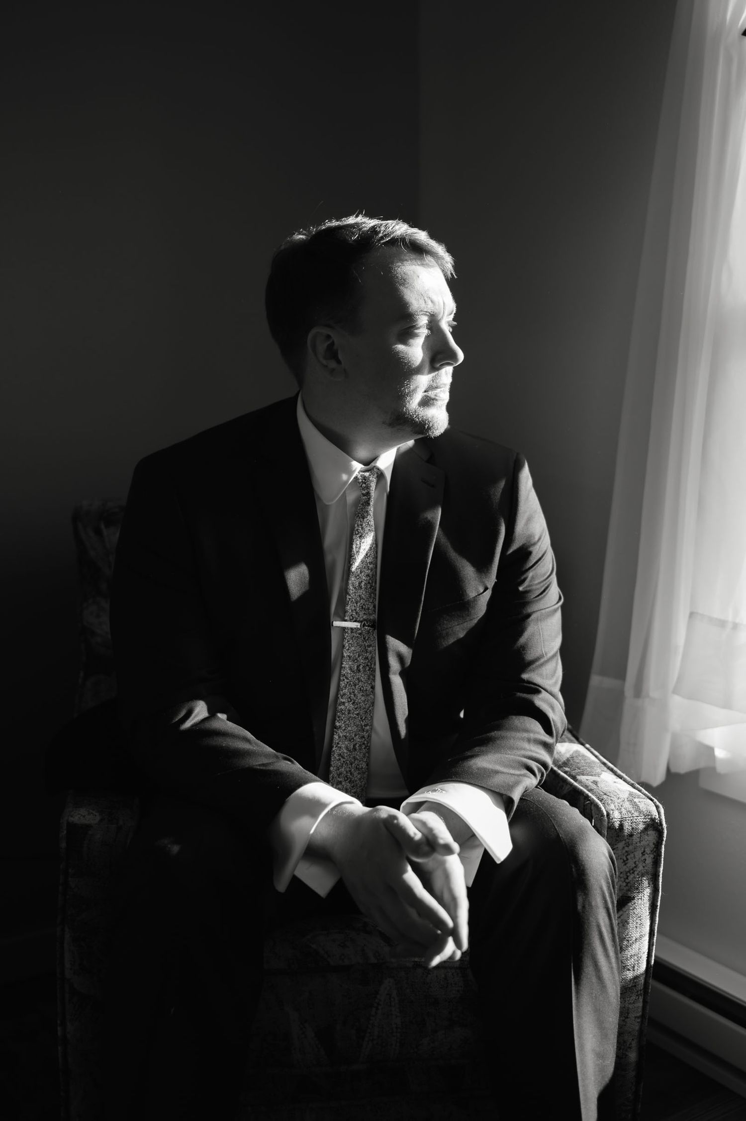 Dapper groom in dark suit and tie sits thoughtfully by a window in a black and white portrait.