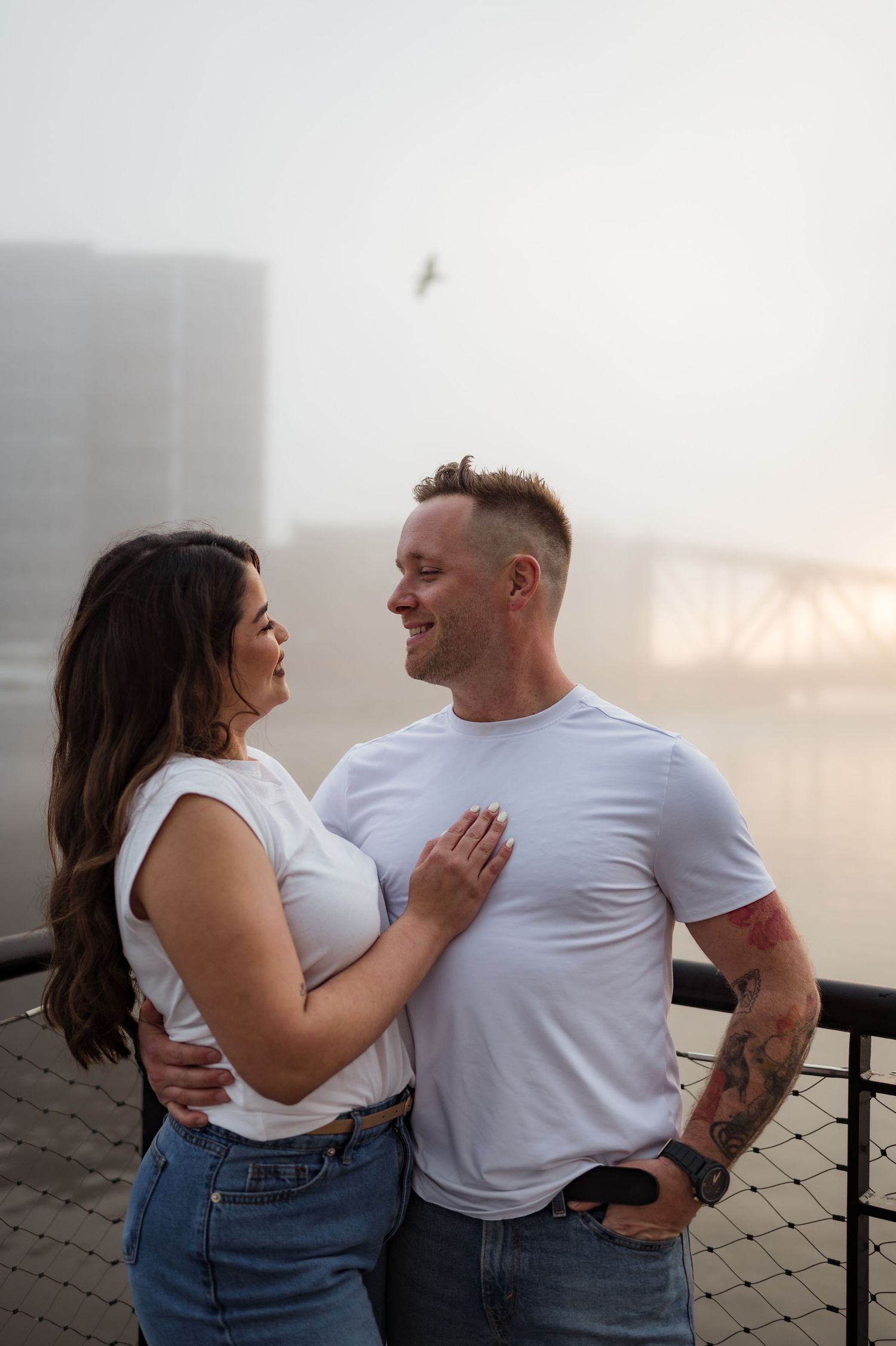 Couple sharing a romantic moment together on a foggy pier by the water, with bridge visible in background.