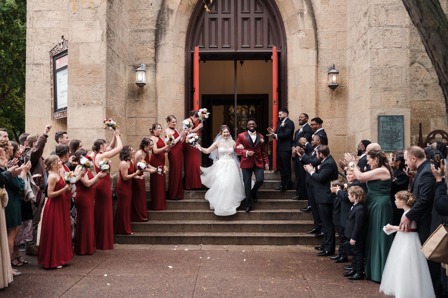 A bride and groom exit a stone church surrounded by cheering bridesmaids in red dresses and groomsmen.