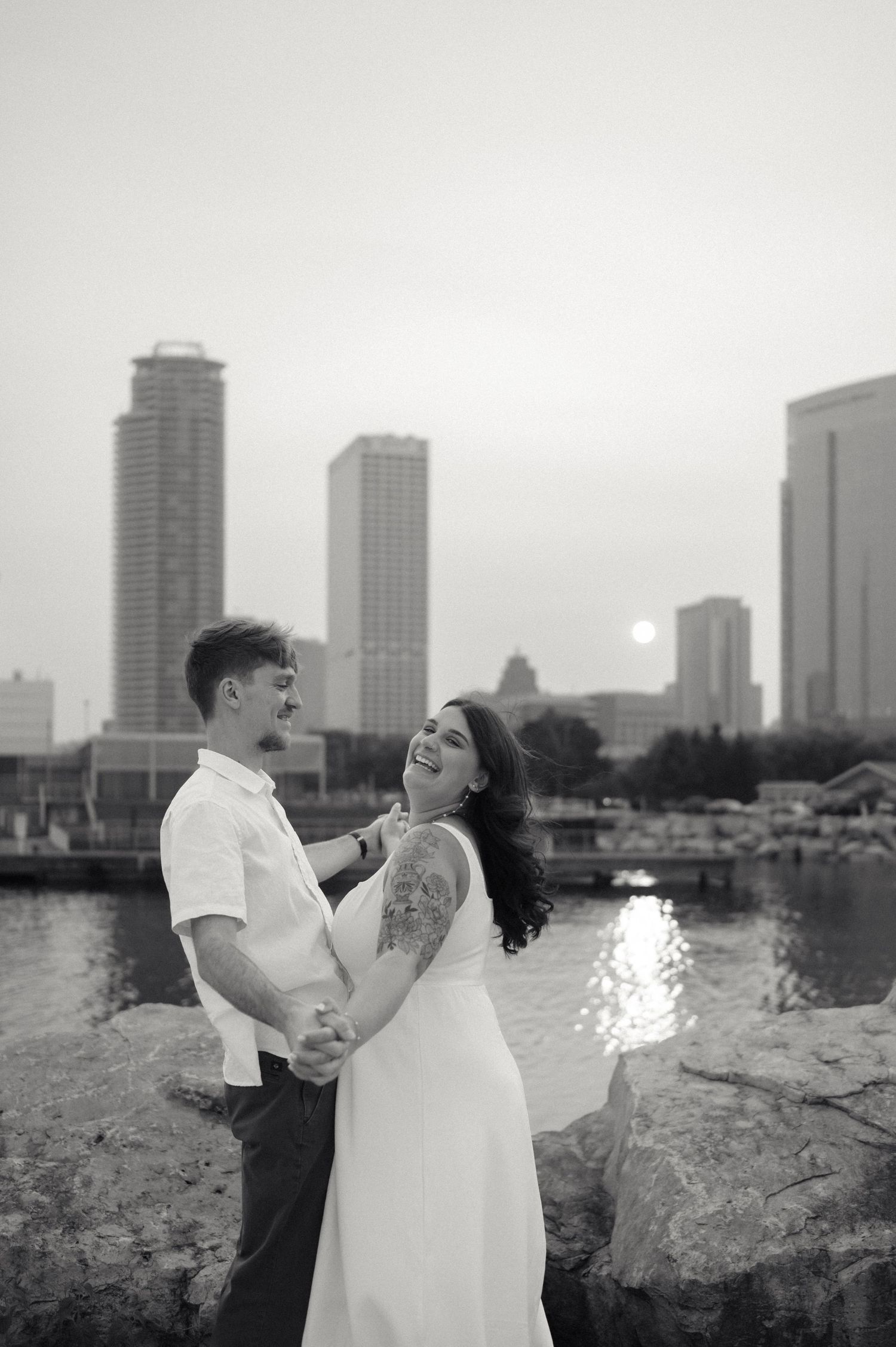 A romantic couple embraces on rocks by a waterfront with city skyscrapers and sunset in the background.