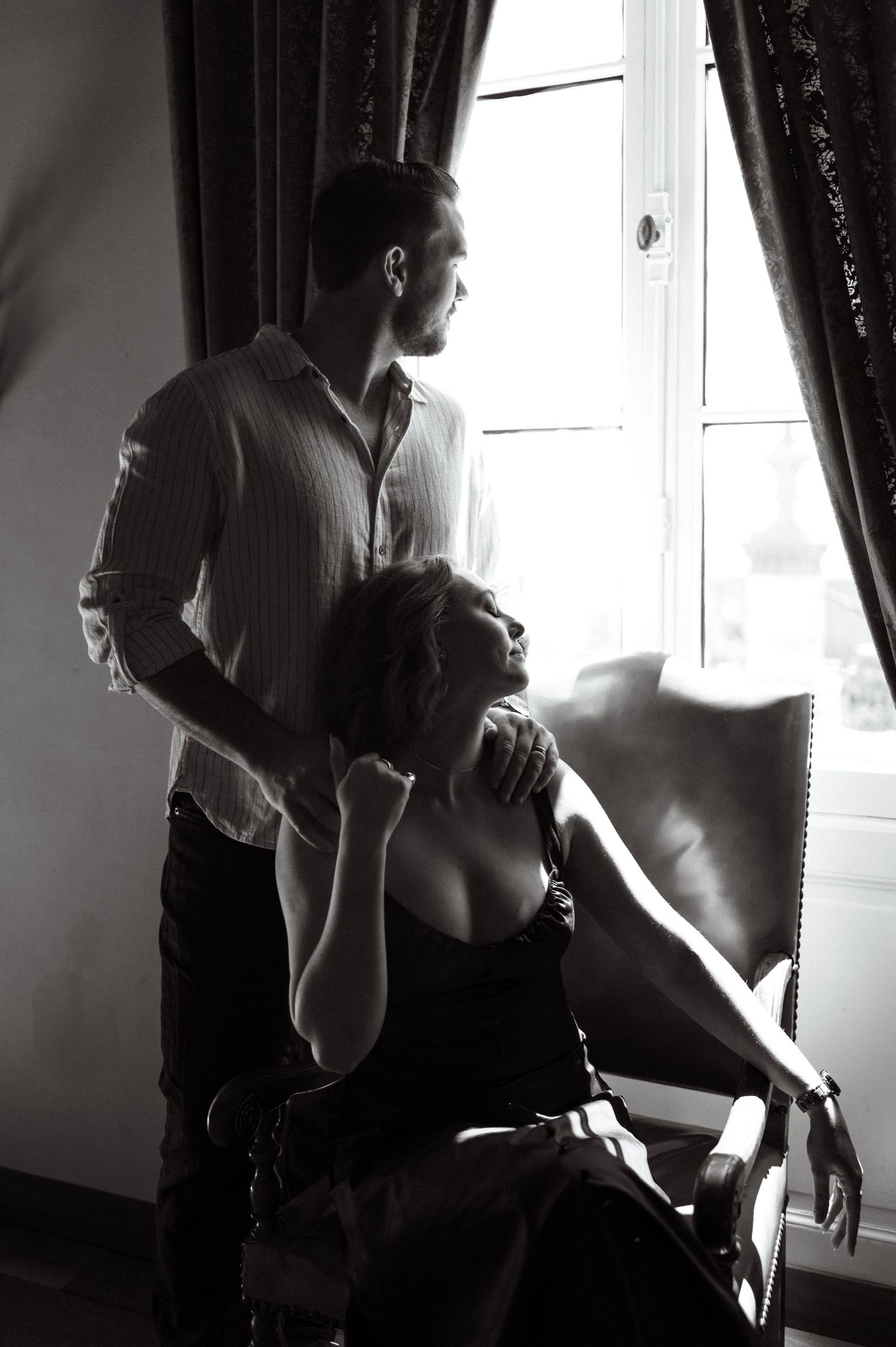 Romantic black and white portrait of couple near window, woman seated in chair looking up at man.