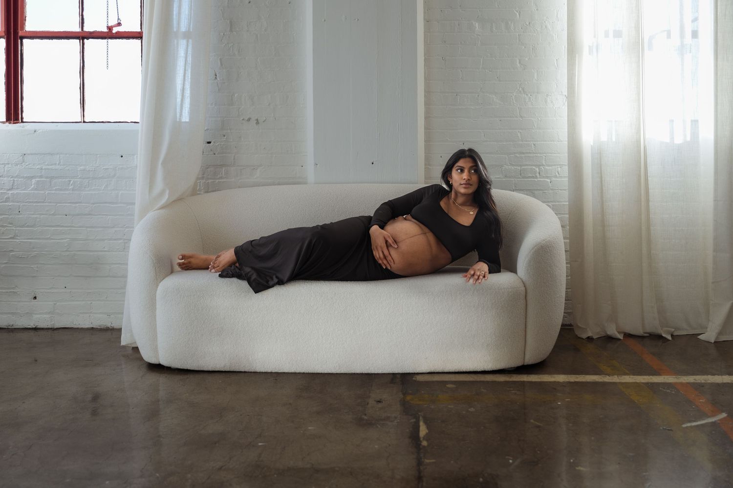 Pregnant woman in black dress reclines on white curved sofa in bright studio.