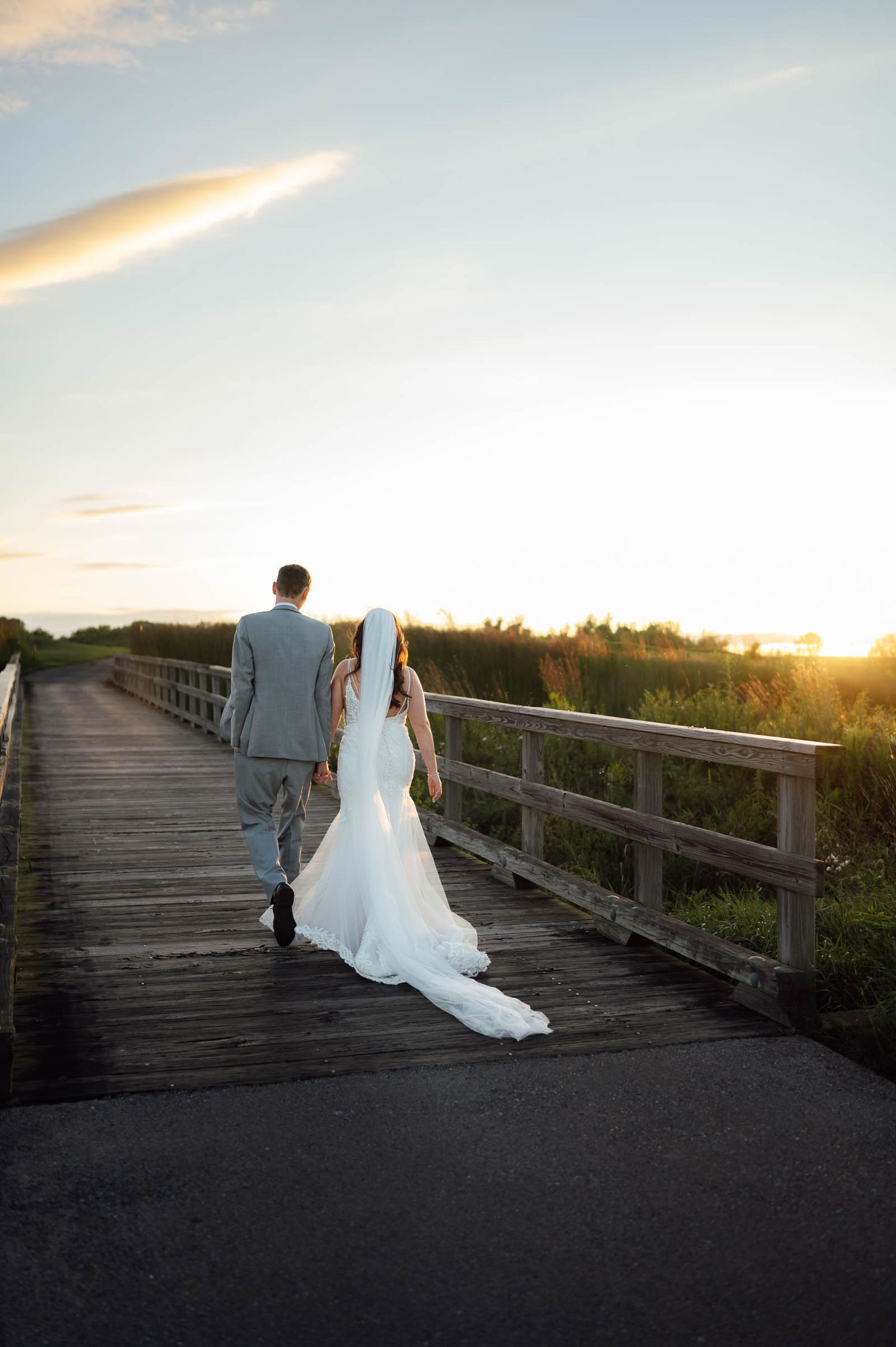 Bride and groom walk hand in hand along a wooden boardwalk toward a golden sunset.