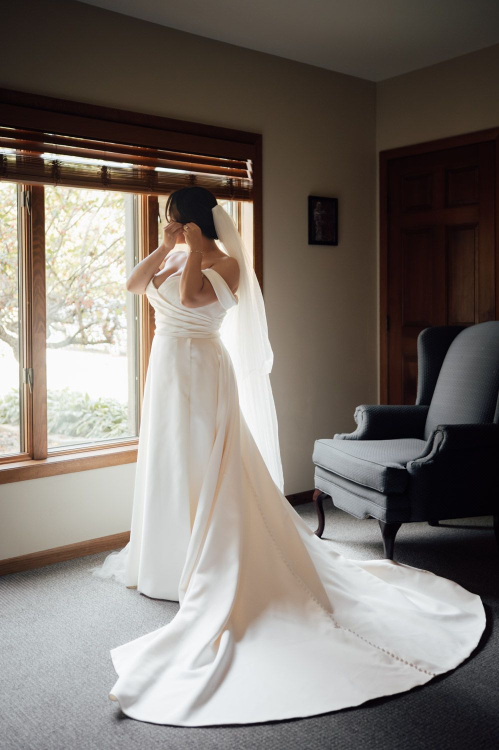 Bride in elegant white off-shoulder gown with long train and veil standing by window.