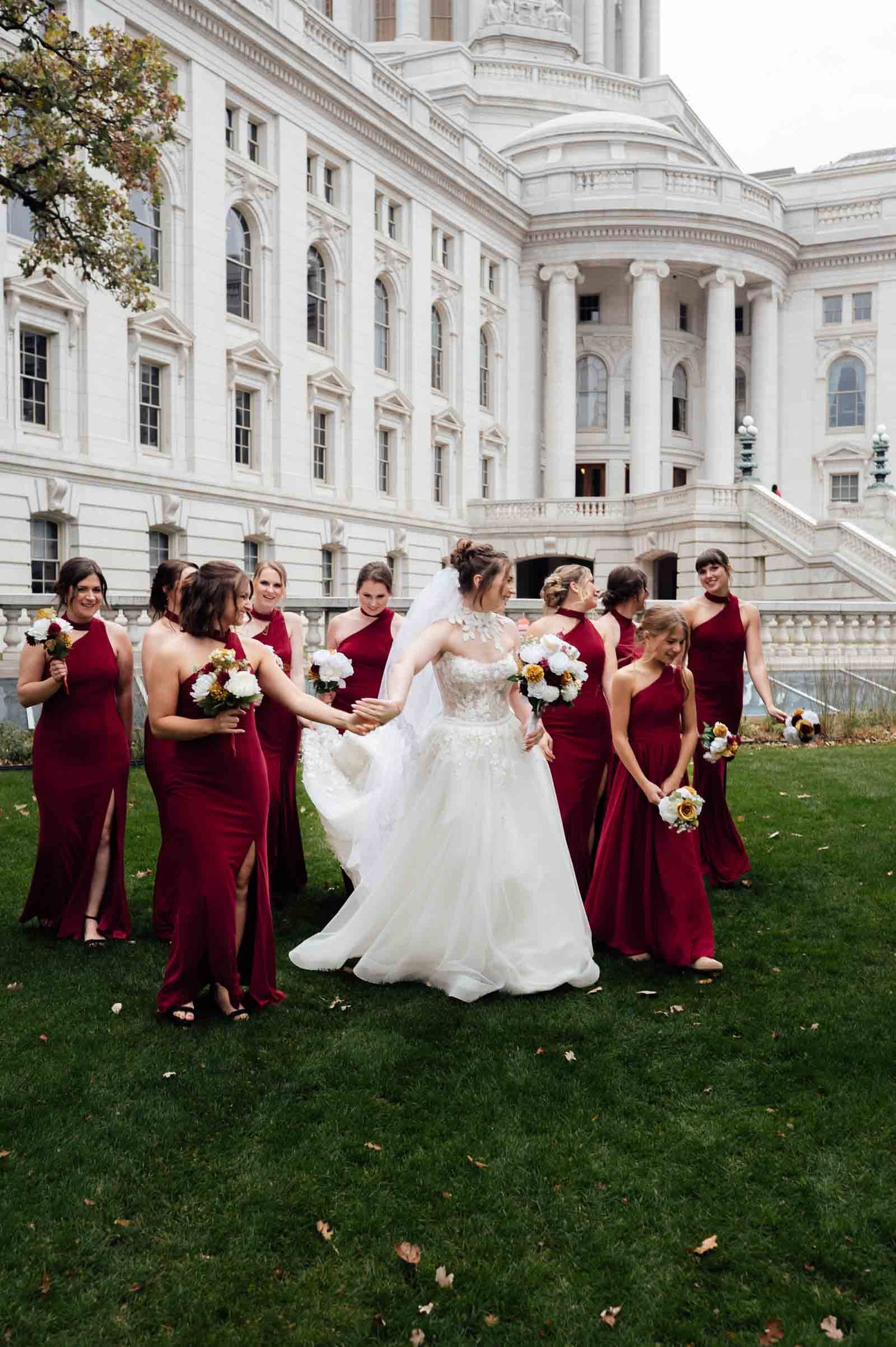 Bride in white gown celebrates with bridesmaids in burgundy dresses outside a grand white capitol building.