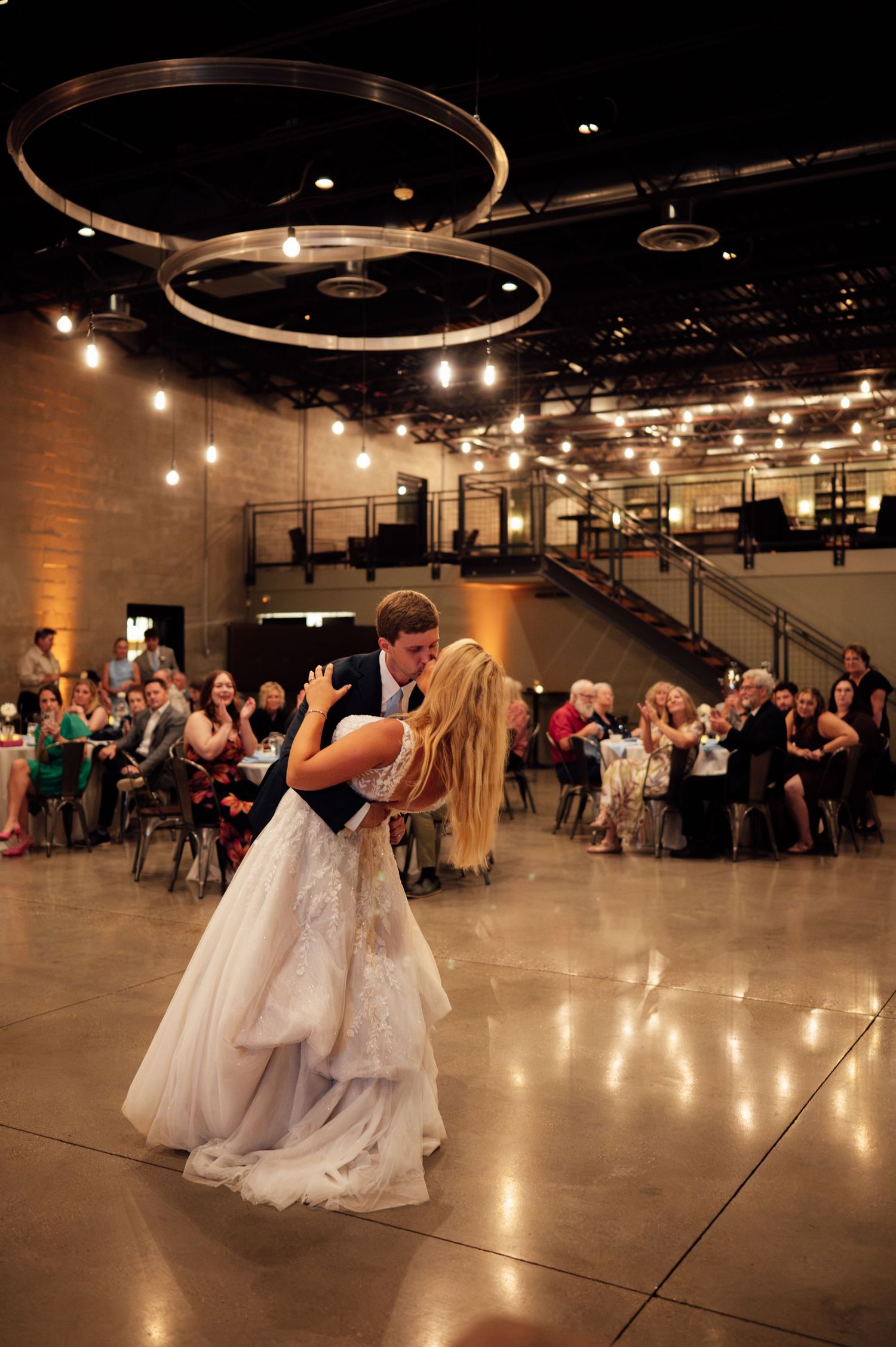 Bride and groom share first dance dip at elegant wedding reception with string lights and circular chandeliers.