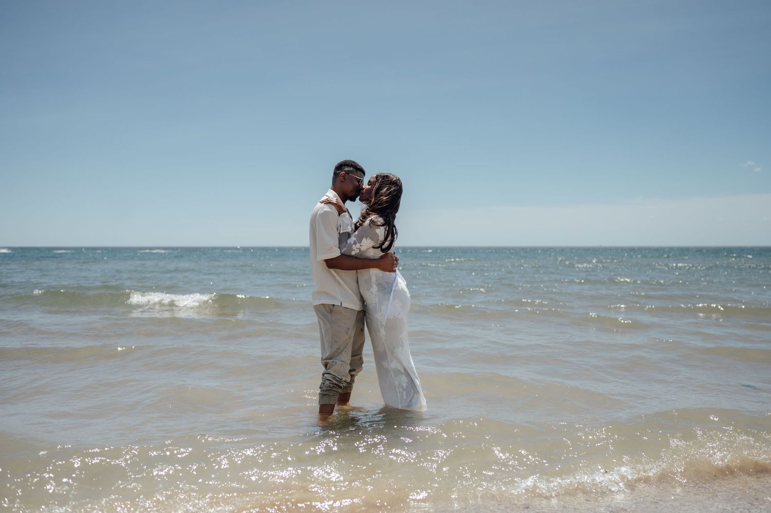 Couple embracing romantically in ocean waves on a sunny beach, wearing white and beige outfits.