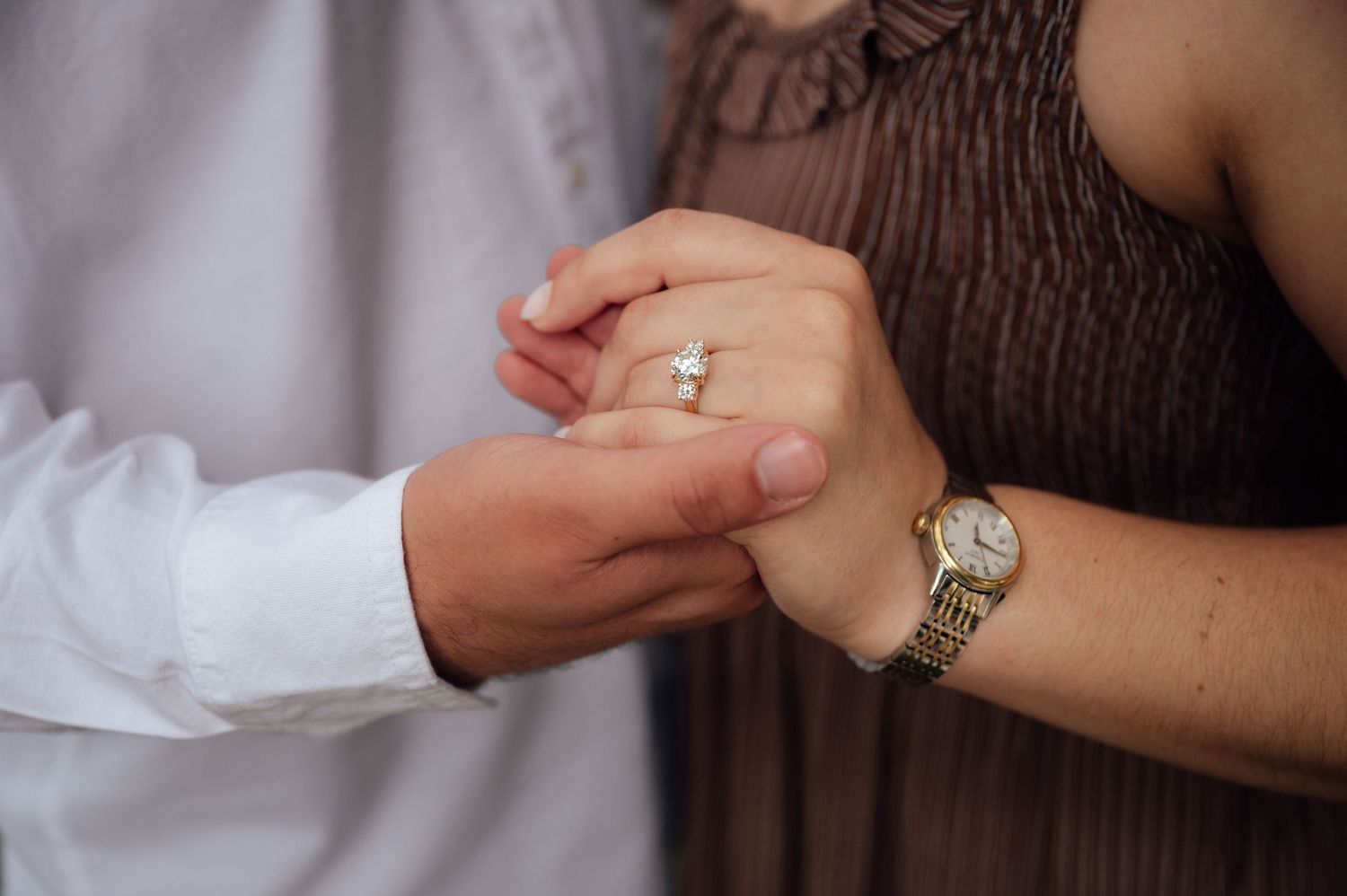 Close-up of couple holding hands, woman wearing diamond engagement ring and gold watch, man in white shirt.