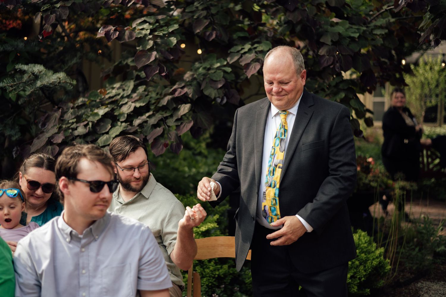 Man in floral tie and suit smiles at outdoor garden wedding reception with seated guests.