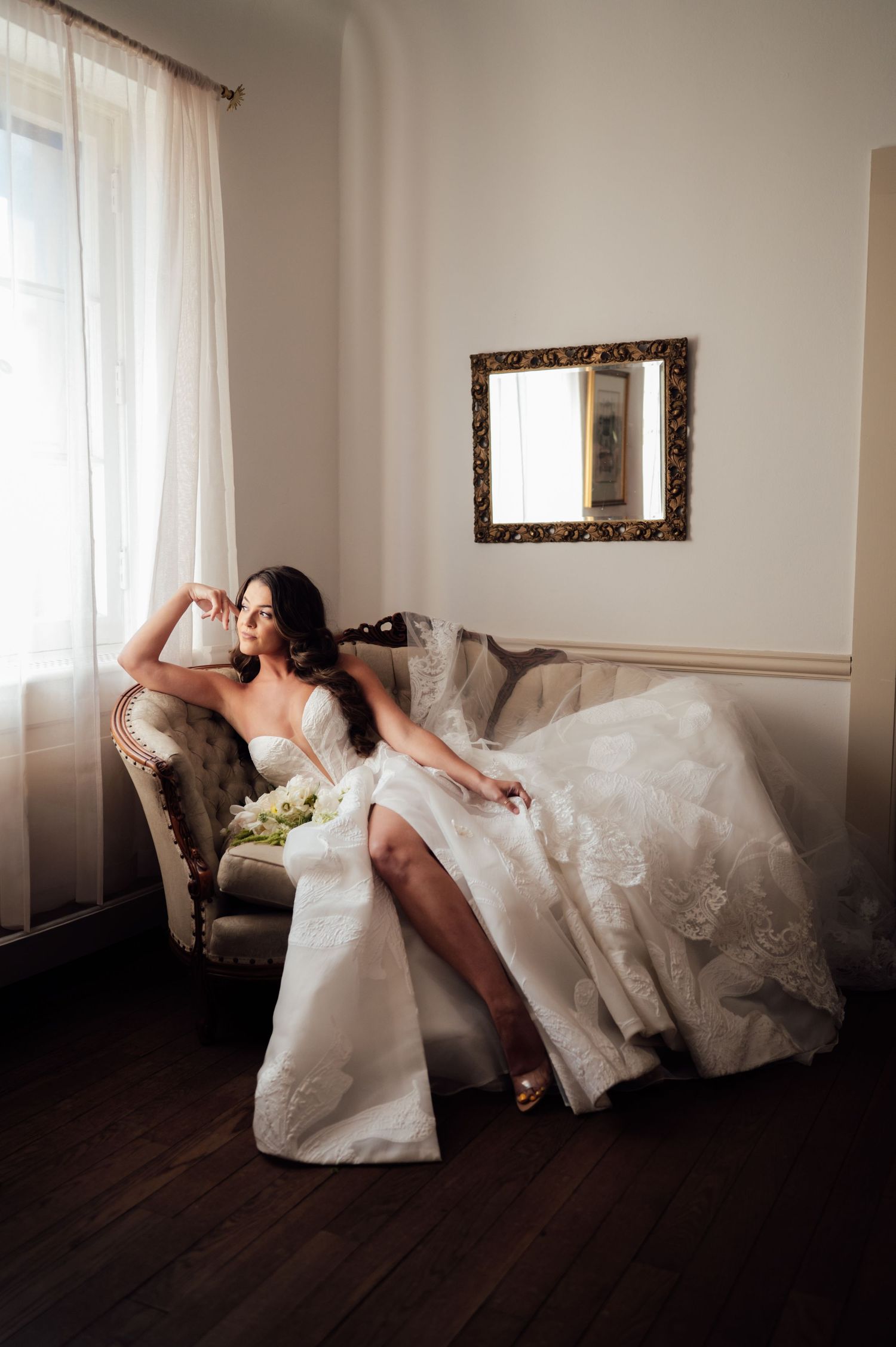 Bride in strapless lace wedding gown reclining elegantly on vintage sofa by window with sheer curtains.