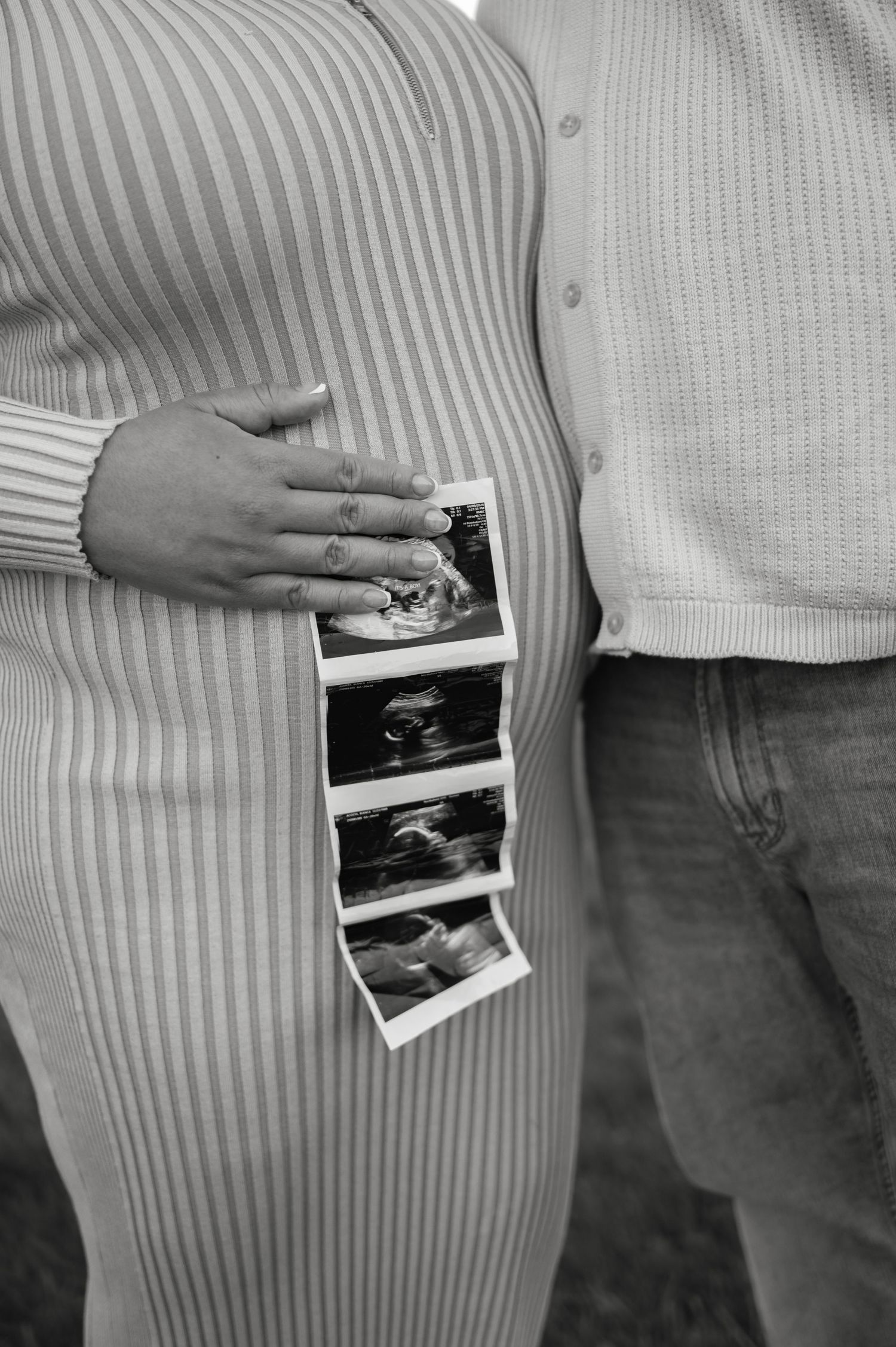 Black and white photo of pregnant woman holding ultrasound photos beside her partner outdoors.