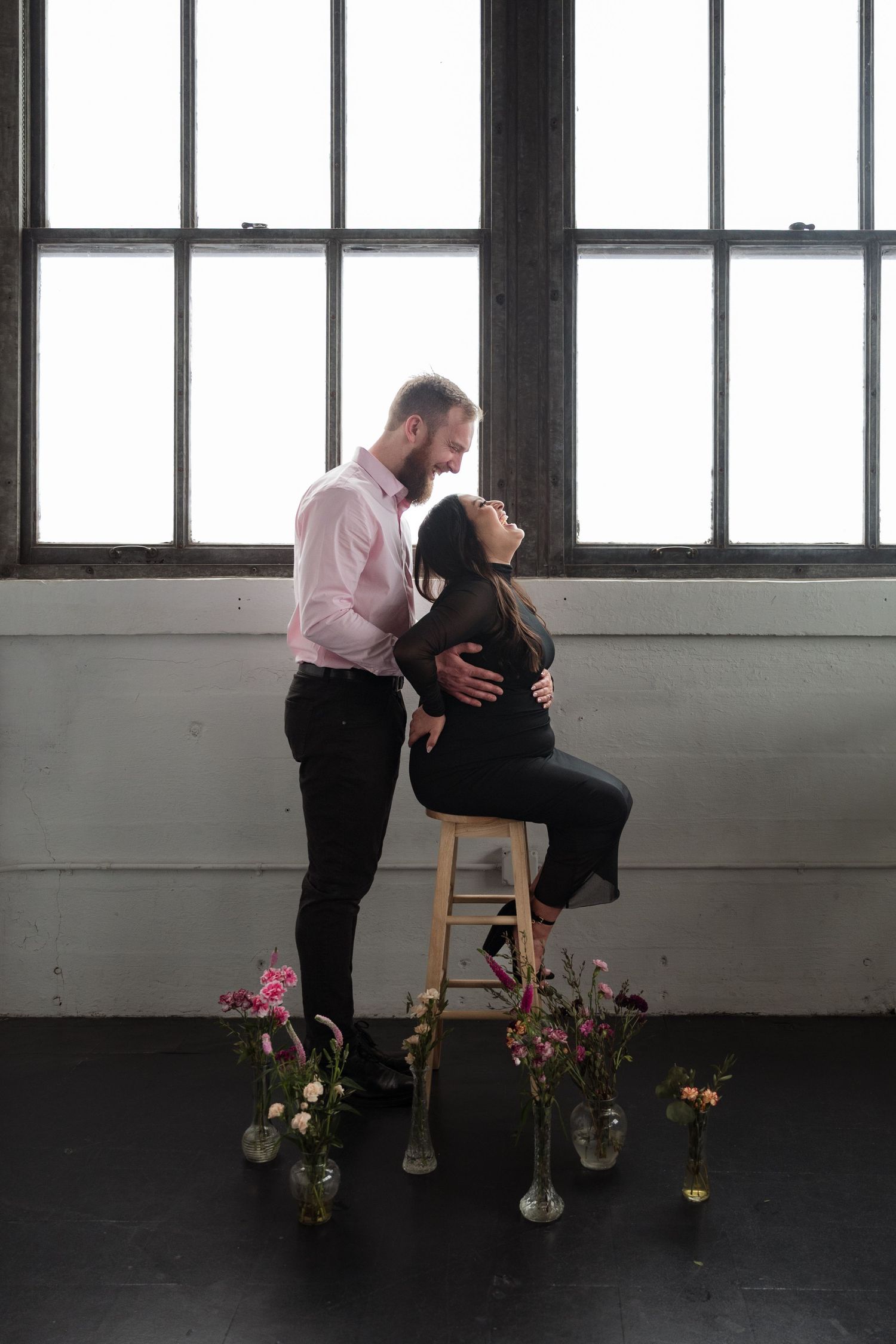 Couple embracing on stool surrounded by flowers in front of large windows.