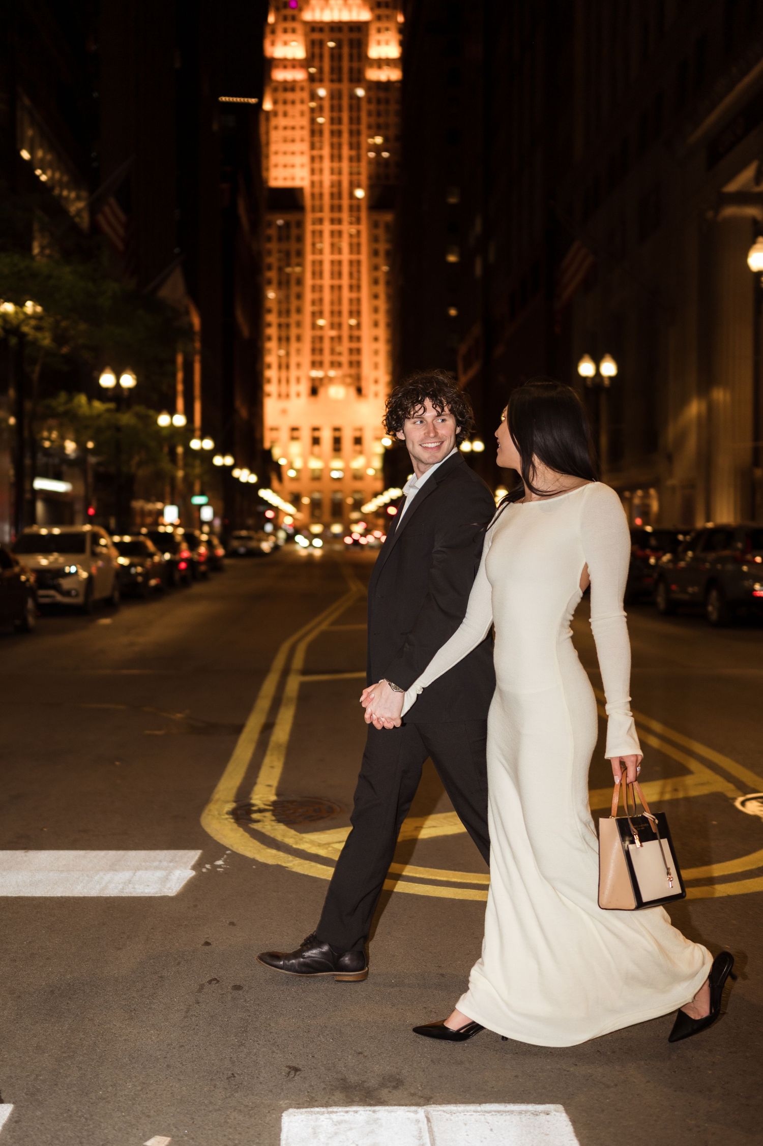 Newlywed couple crossing a Chicago street at night with a glowing skyscraper illuminated in the background.