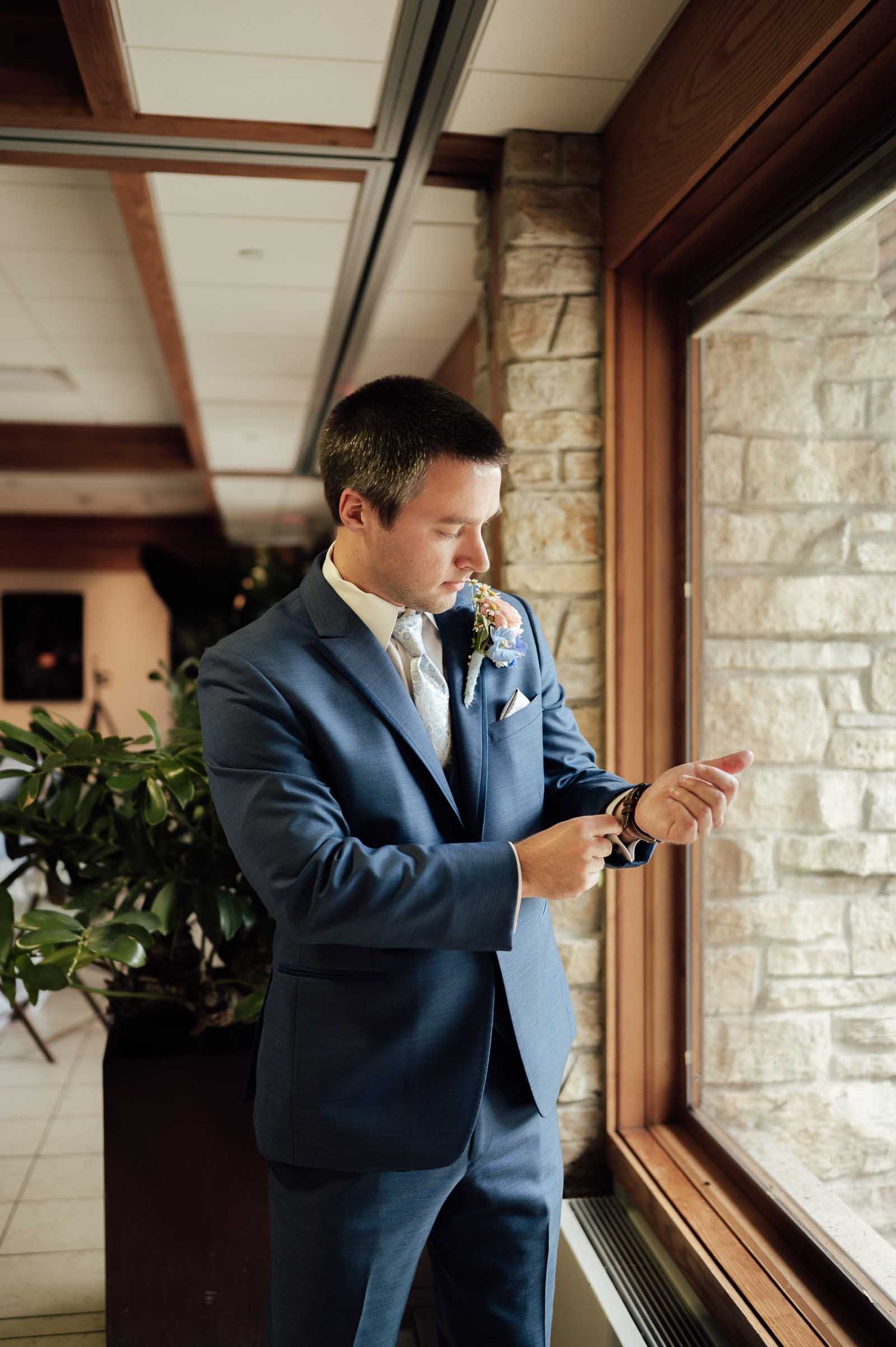Groom in navy suit checking watch by stone wall window before wedding ceremony.