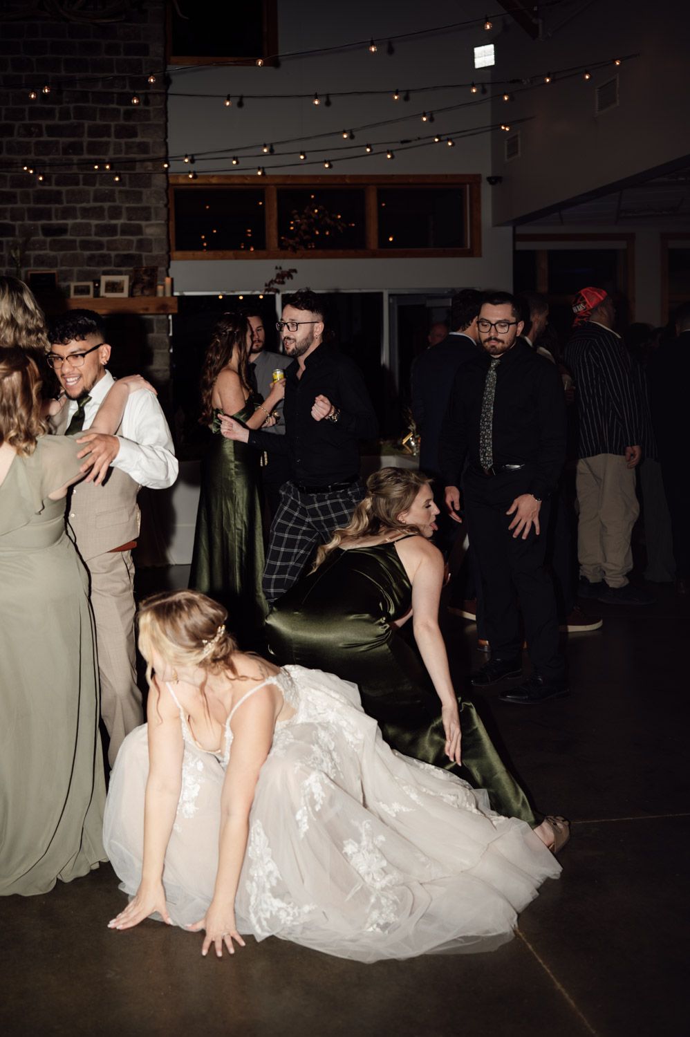 Bride dancing energetically with guests at lively indoor wedding reception with string lights.
