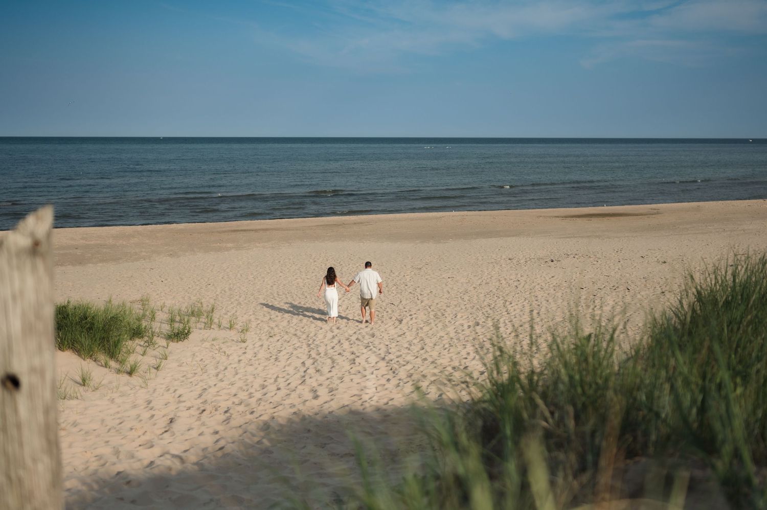 A tranquil sandy beach shoreline stretches along a calm blue ocean under clear summer skies with dune grass in view.