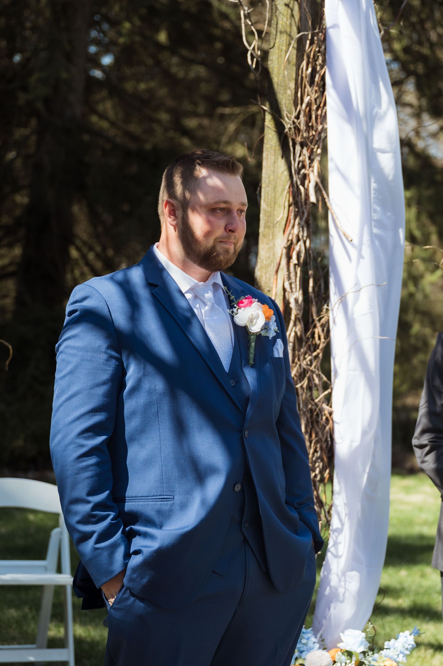 A wedding attendee in a stylish navy blue suit with pink boutonniere stands near a white fabric-draped ceremony arch outdoors.