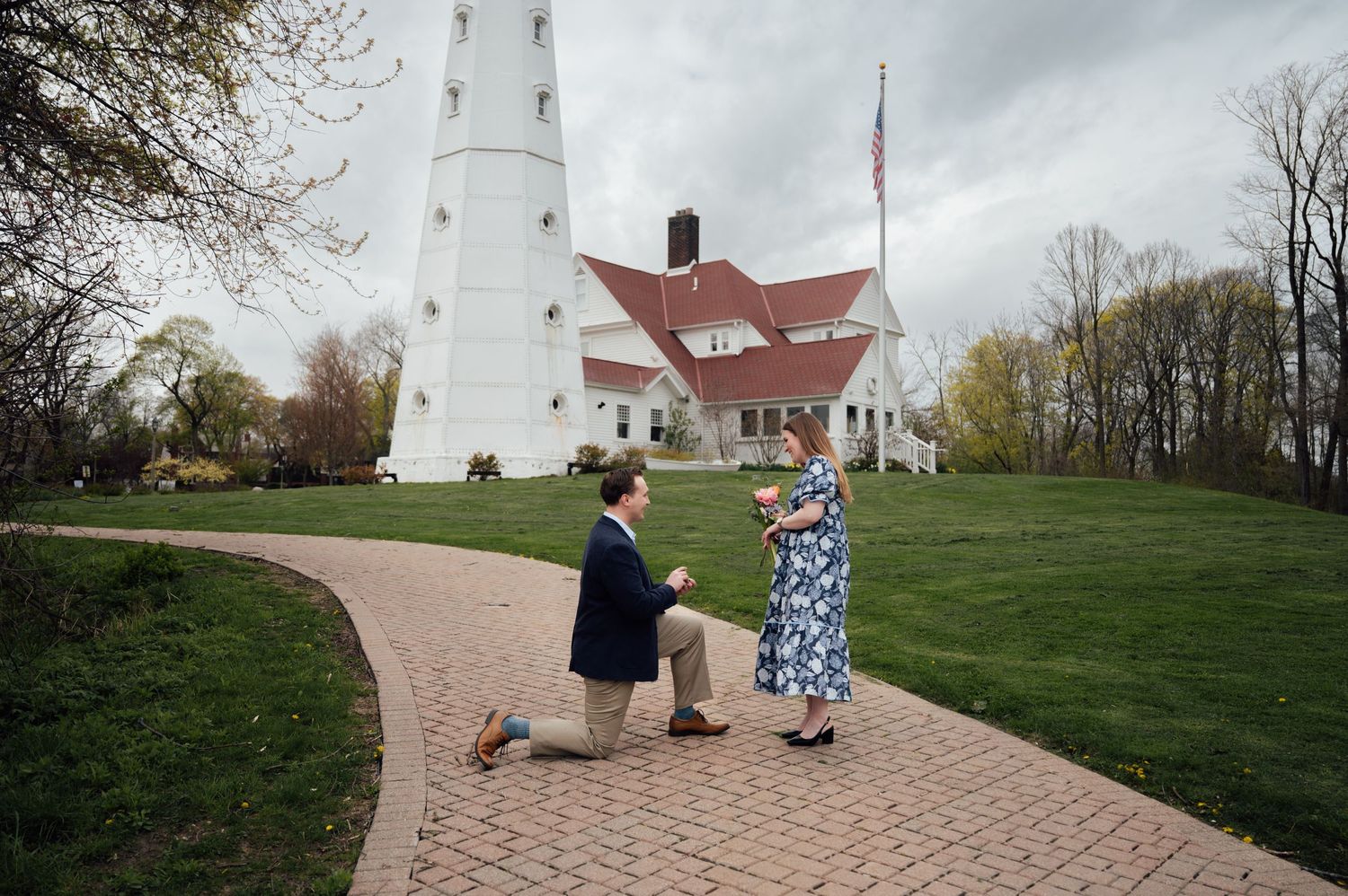 Man proposing on one knee to woman on brick path near white lighthouse on cloudy day.