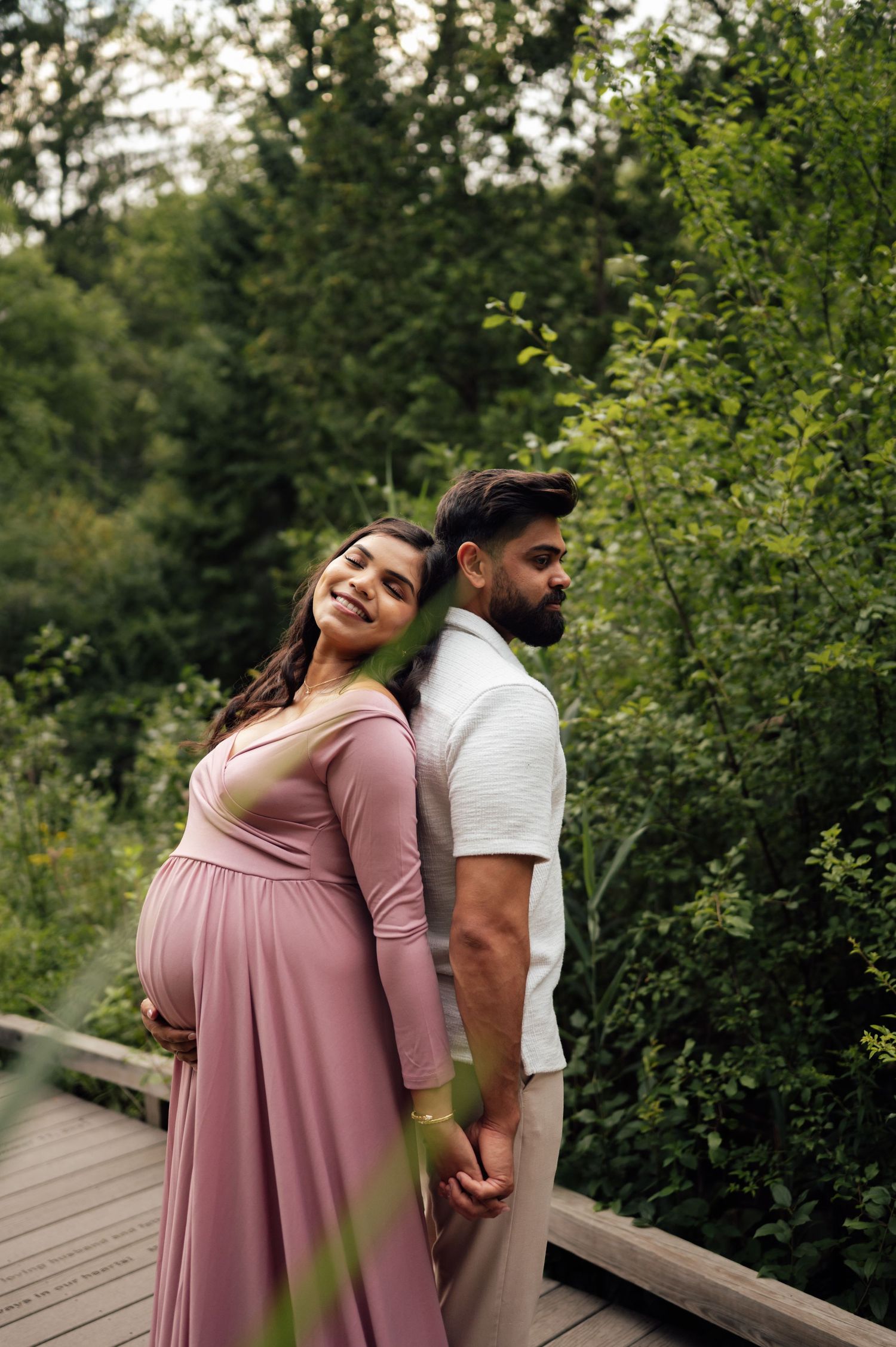 Pregnant woman in pink dress holds hands with man in white shirt on wooden bridge surrounded by lush green trees.