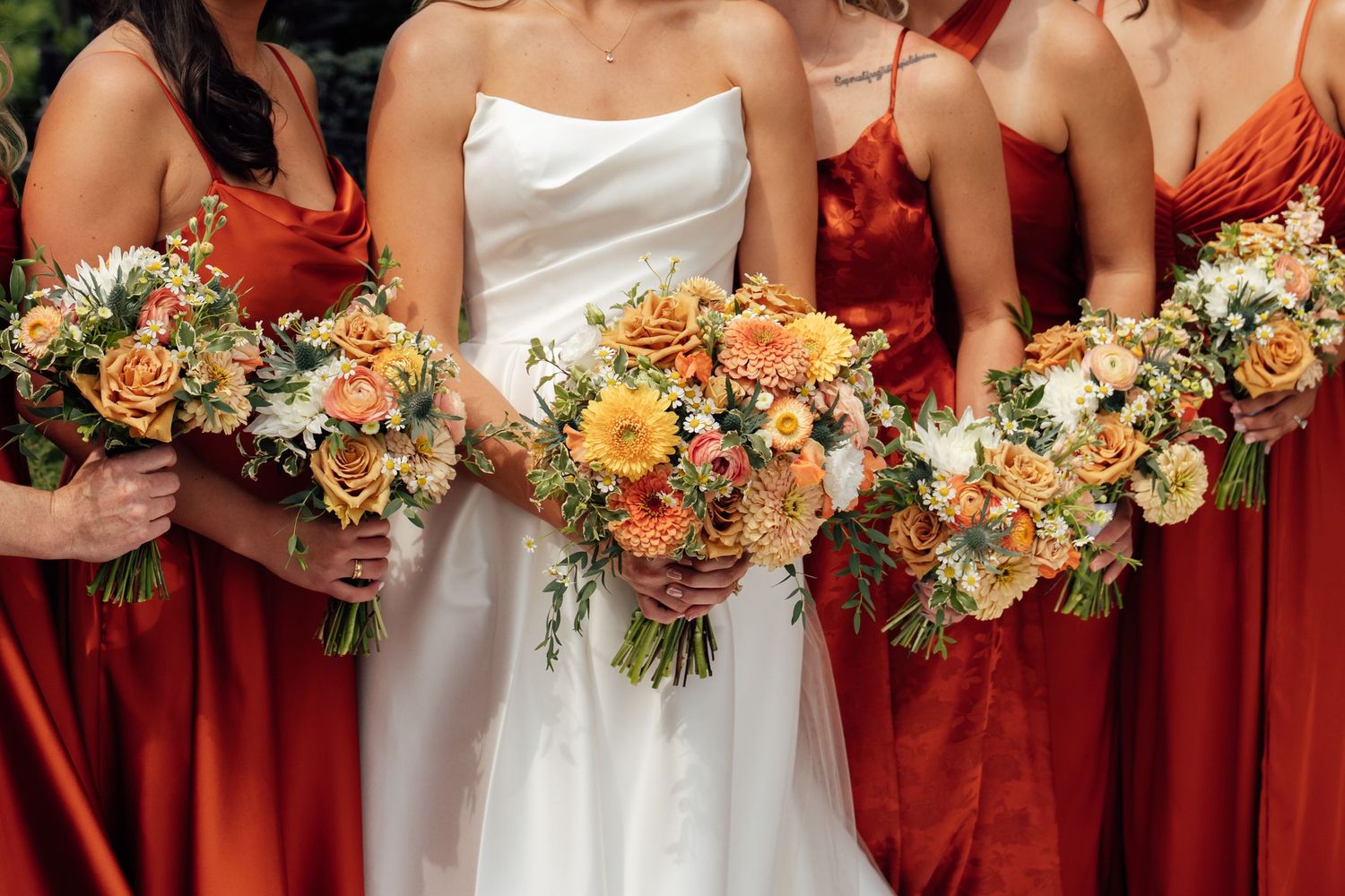 Bride in white gown with bridesmaids in rust orange dresses holding bouquets of peach roses and wildflowers.