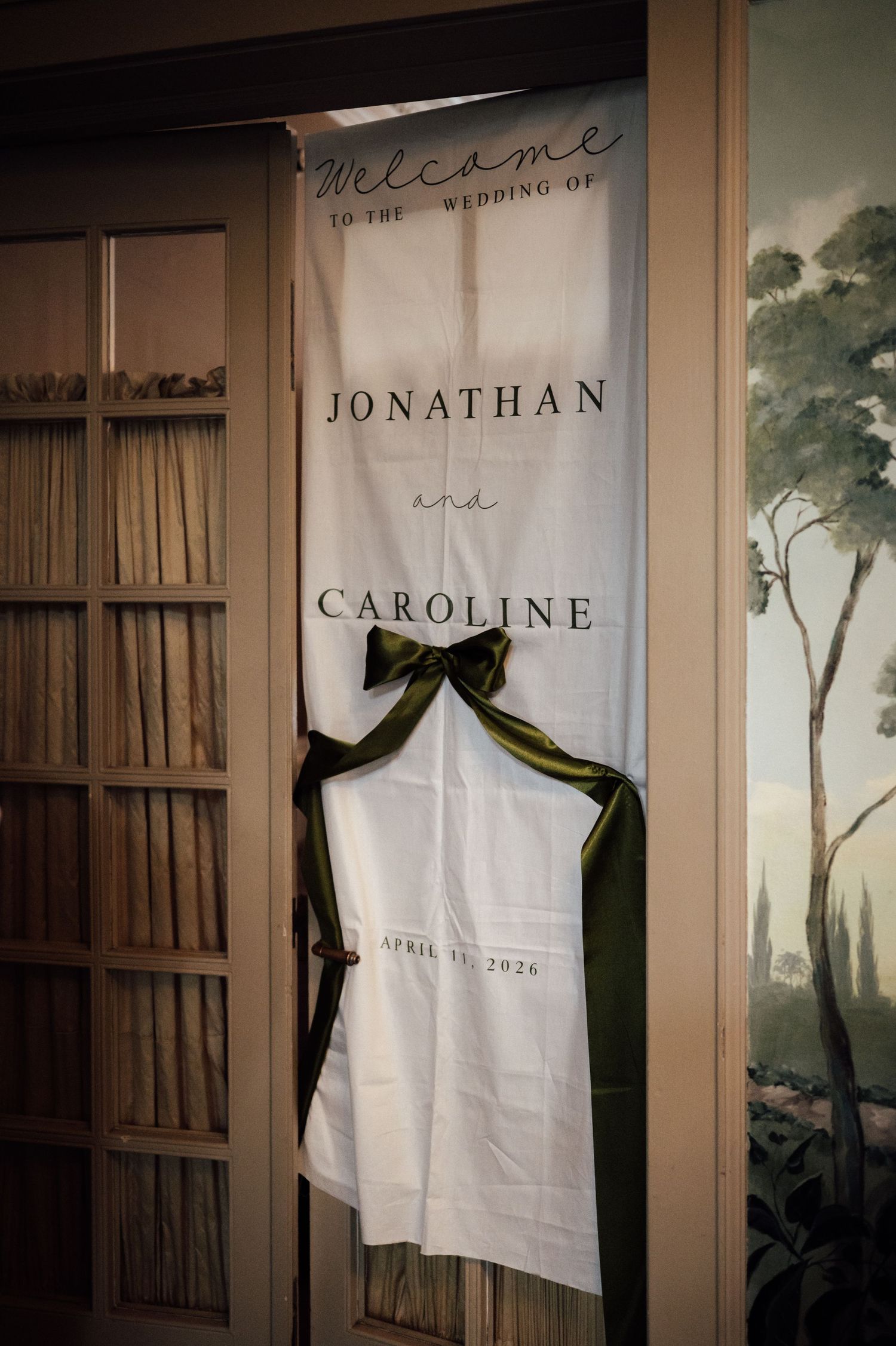 Elegant wedding welcome banner for Jonathan and Caroline, April 2026, adorned with a green satin bow on wooden doors.