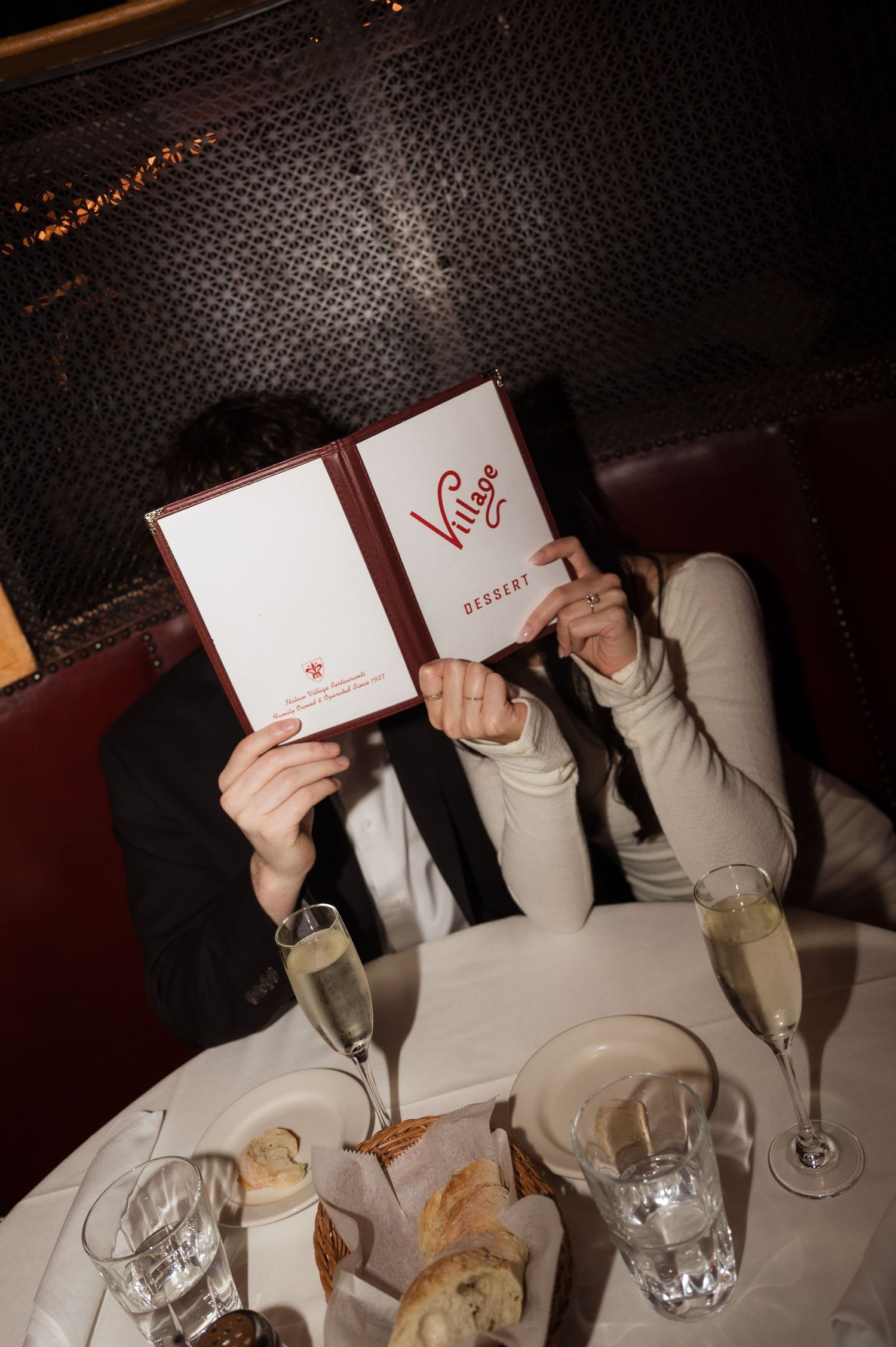 Engaged couple hiding behind a restaurant dessert menu with champagne glasses on the table.