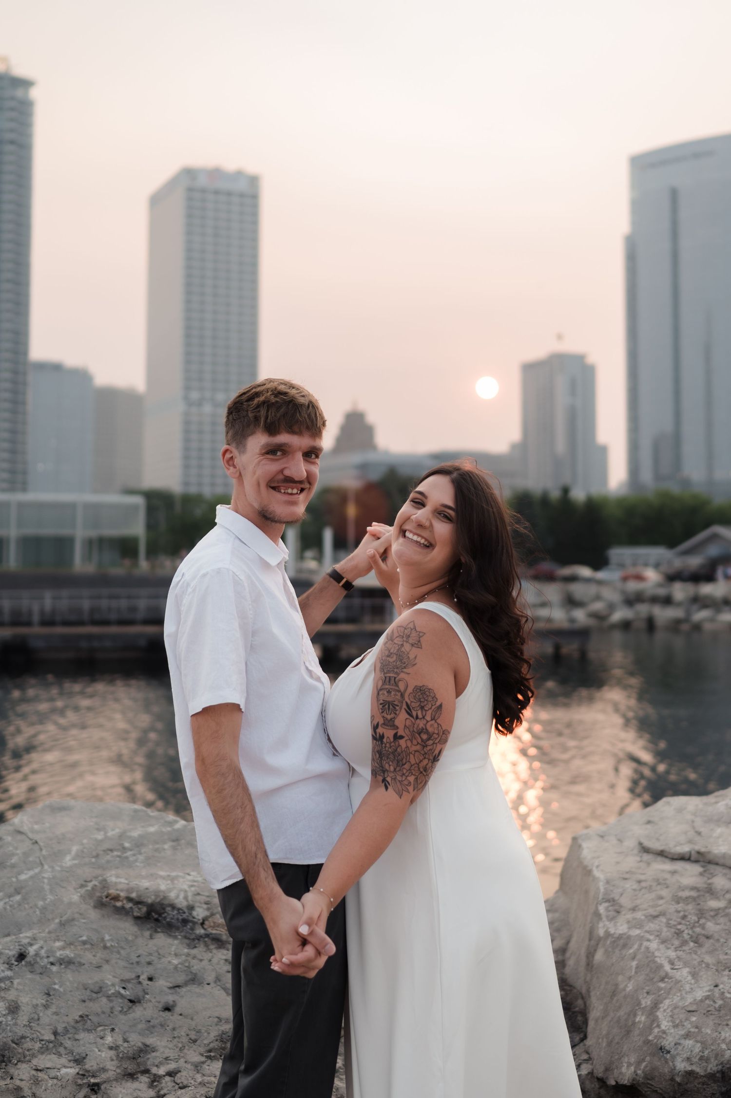 Smiling engaged couple posing at sunset with Milwaukee city skyline reflecting on the water.