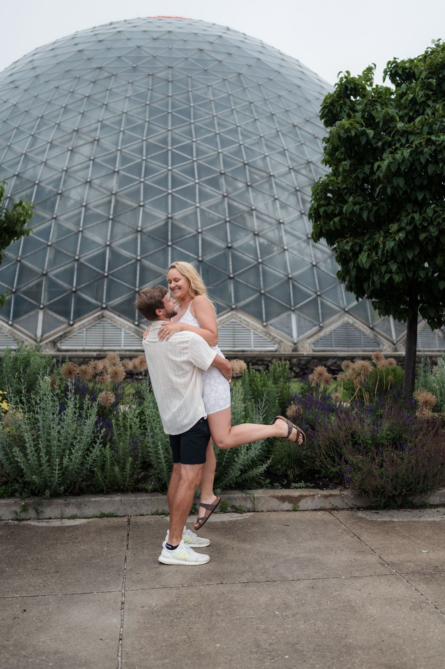 Man joyfully lifting woman in front of a large geodesic dome greenhouse surrounded by gardens.