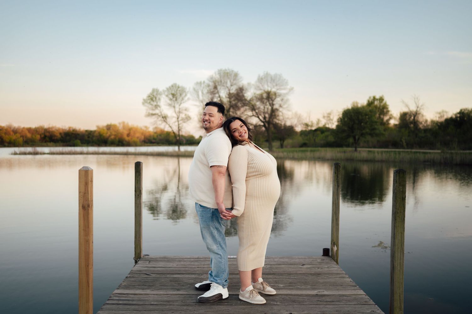 Expecting couple standing back to back on a wooden dock at sunset by a calm lake.