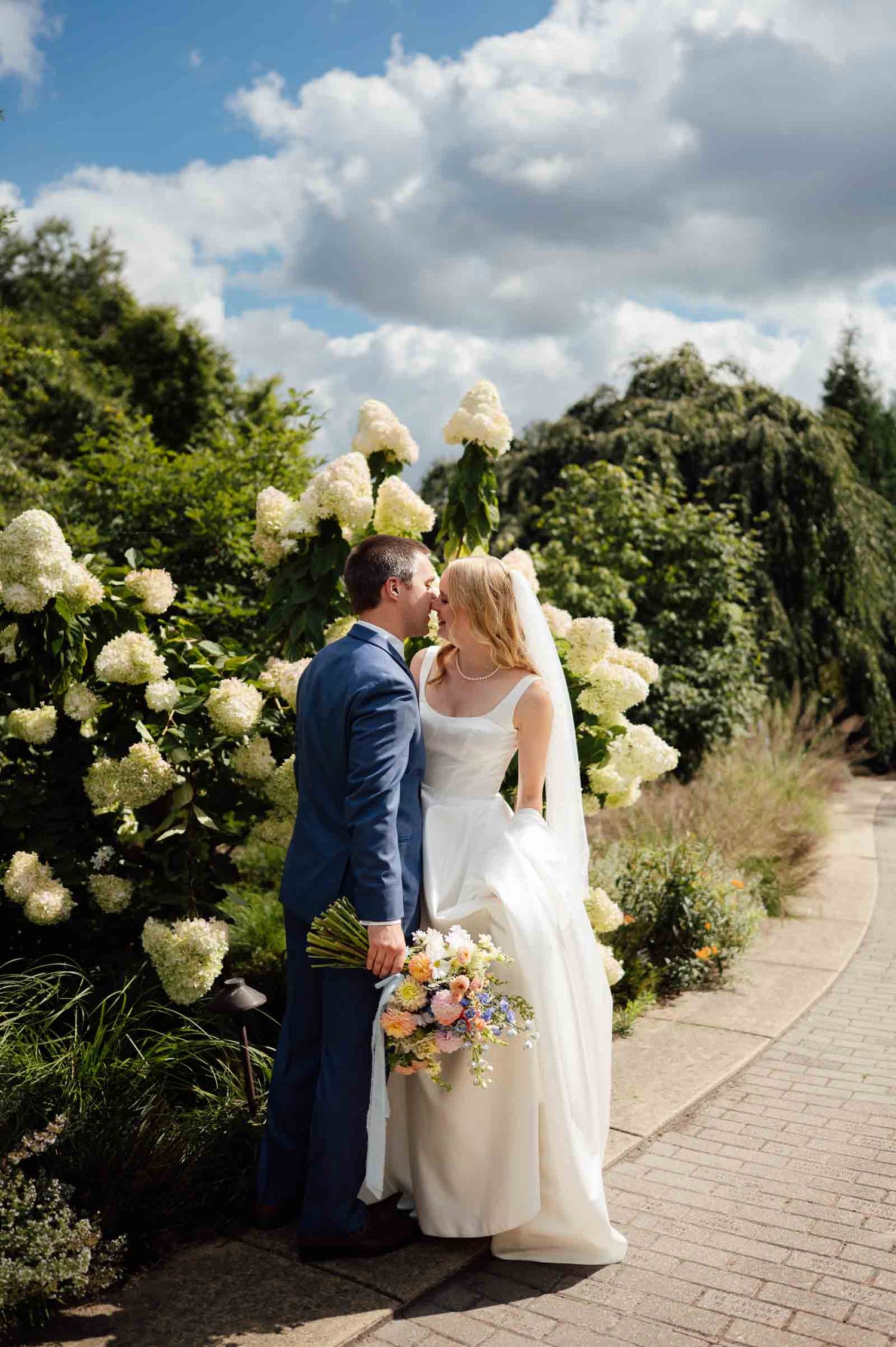 Bride and groom share a kiss beside blooming white hydrangeas on a sunny garden wedding day.