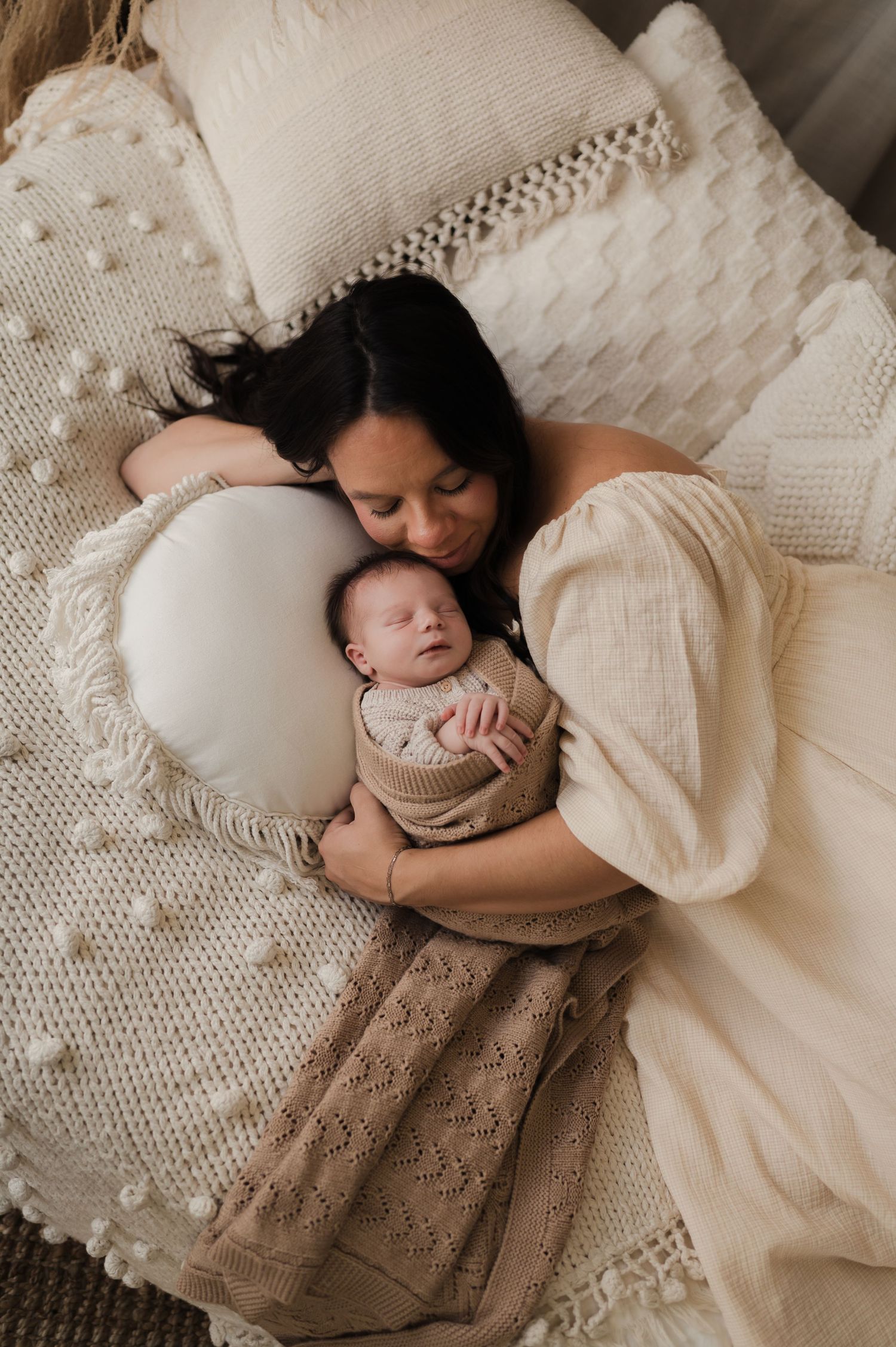 A dark-haired mother in a cream dress cuddles her sleeping swaddled newborn on a cozy textured bed.