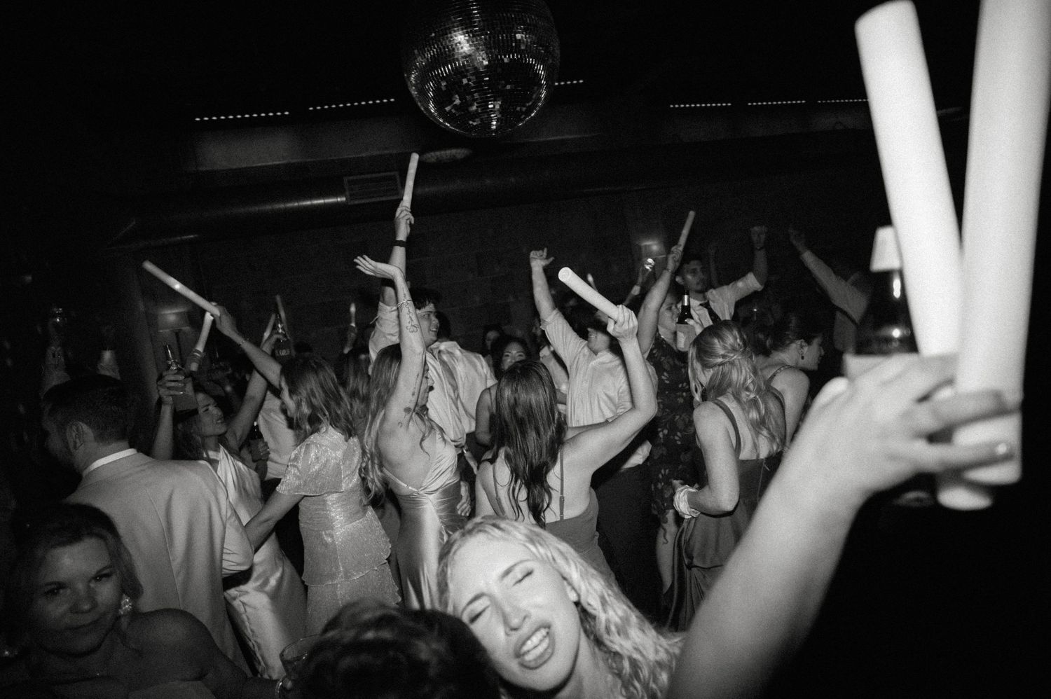 Black and white photo of crowd dancing at a party, holding glow sticks under a disco ball.