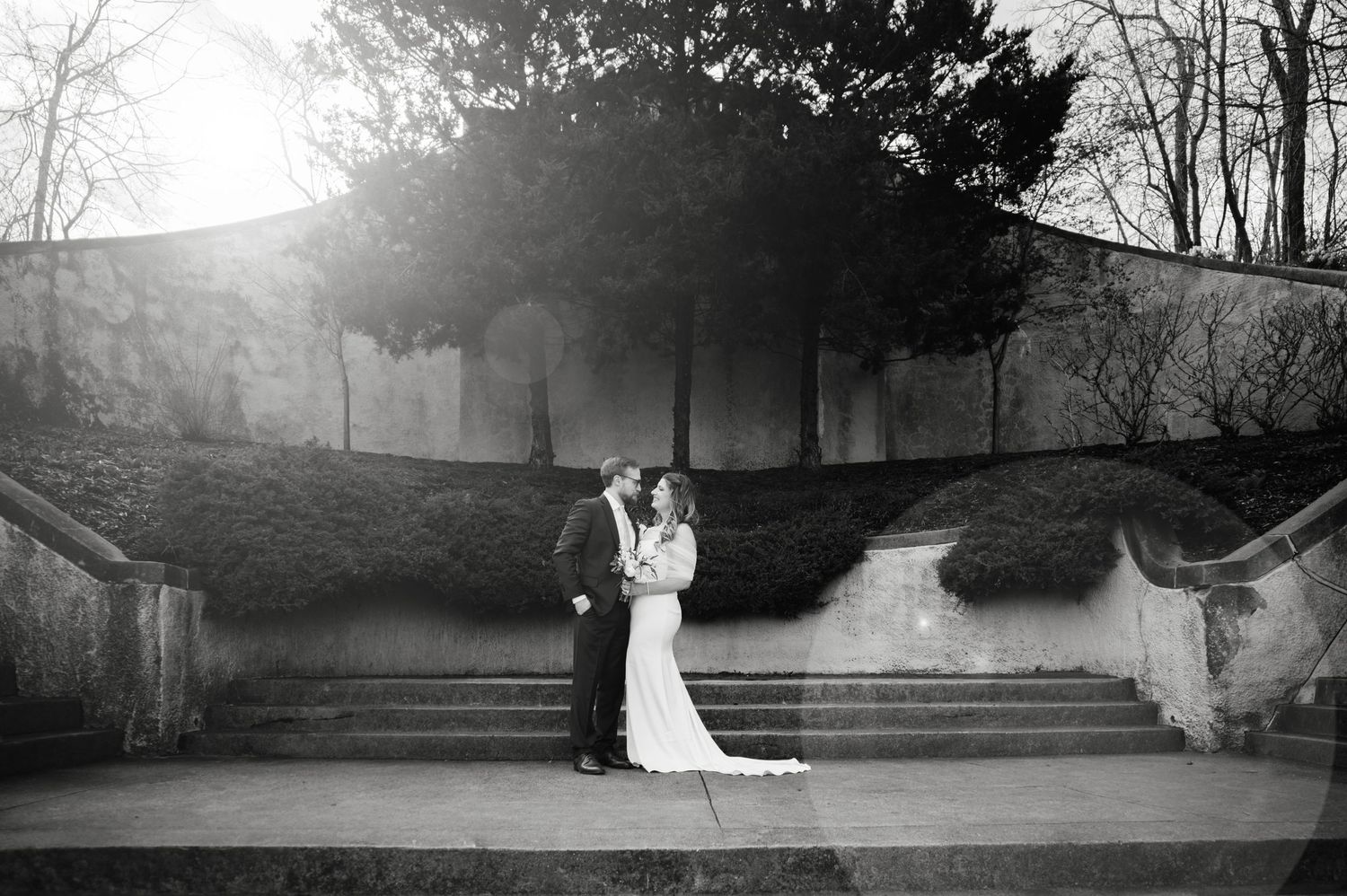 Black and white romantic photo of newlywed couple embracing on stone steps in a garden.