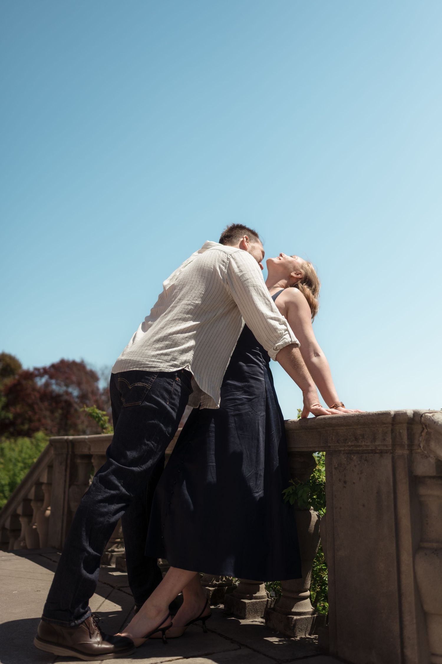Couple sharing a kiss leaning against a stone balustrade under a clear blue sky outdoors.